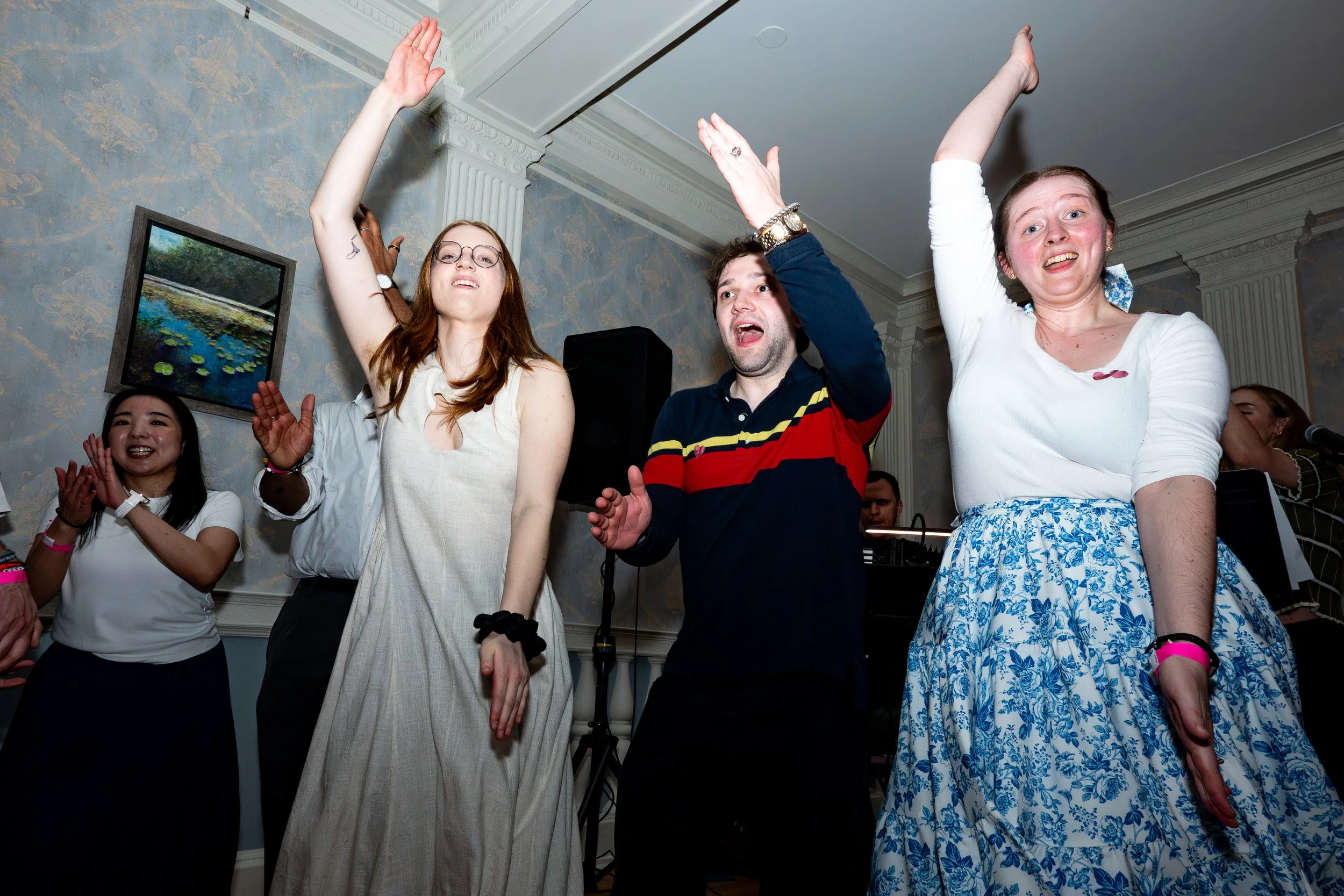 Group of people dancing and celebrating indoors at a party or gathering, with some raising their hands and smiling.
