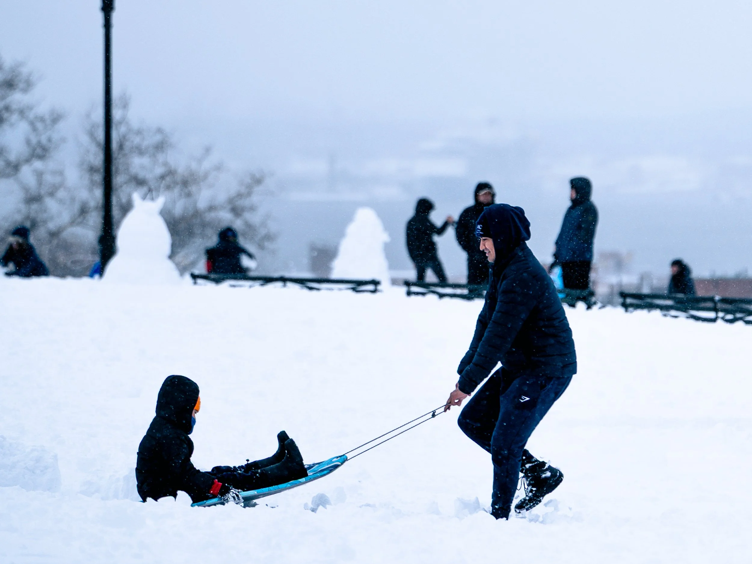 A man pulling a child on a sled in the snow on a winter day, with snowmen and several people in the background.