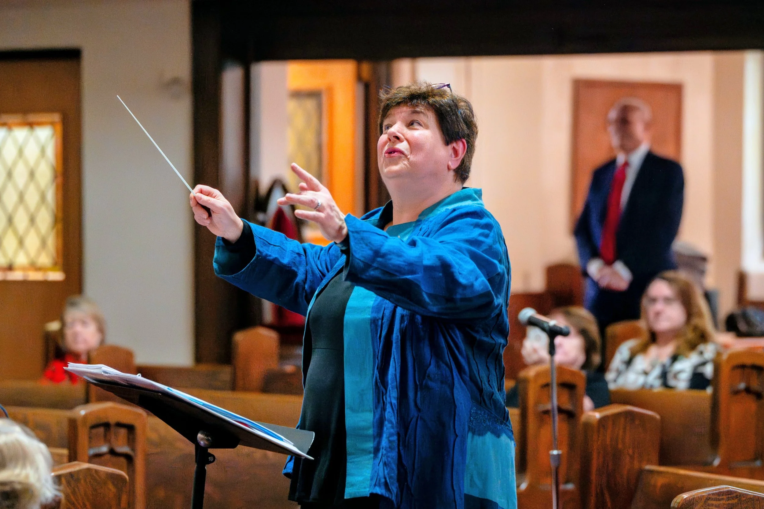 A woman in a blue jacket conducting a choir in a church, with a music stand in front of her and an audience seated behind.