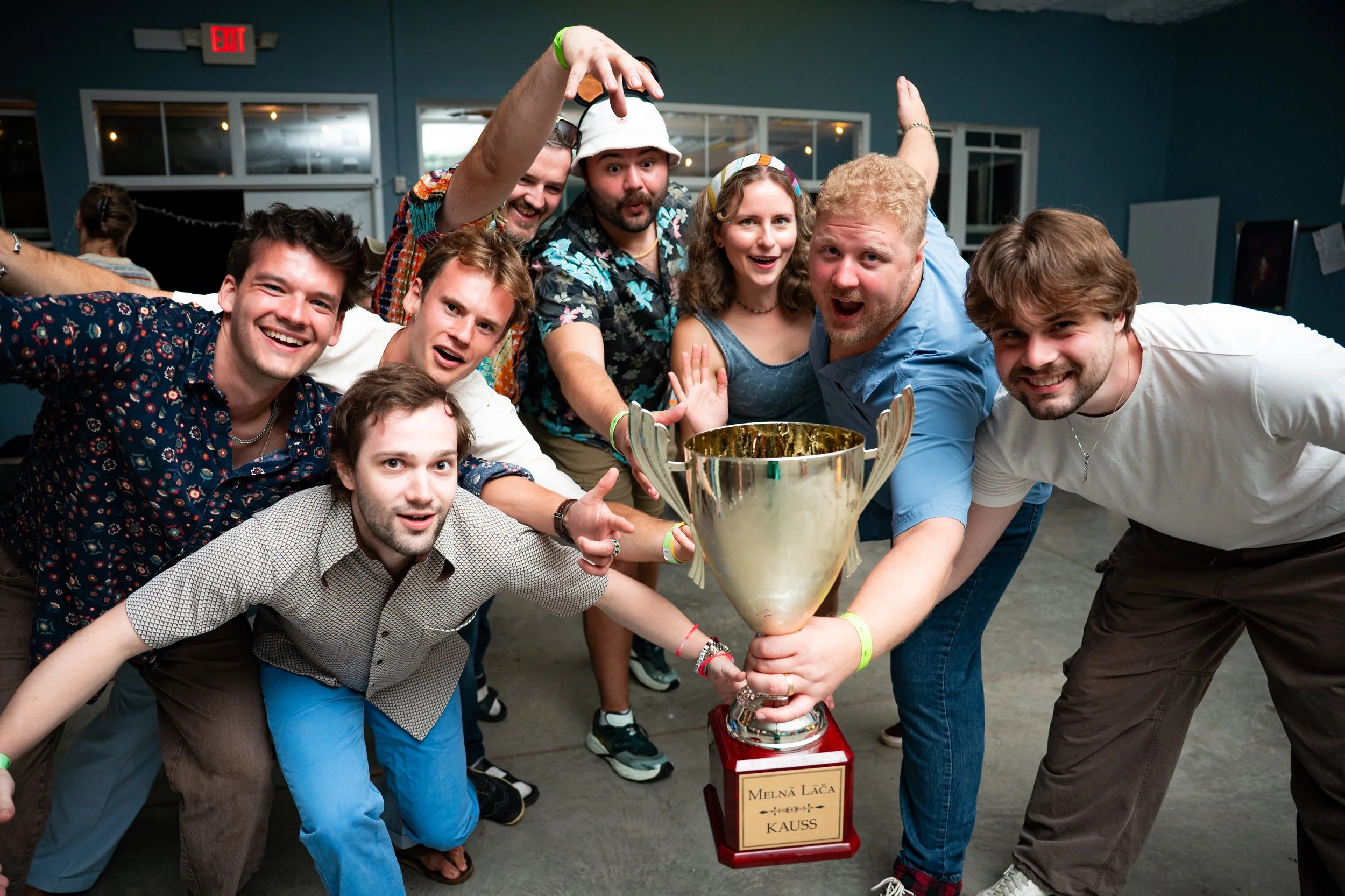 Group of people celebrating a victory around a trophy, with some making celebratory gestures, indoors with blue walls and a red exit sign in the background.