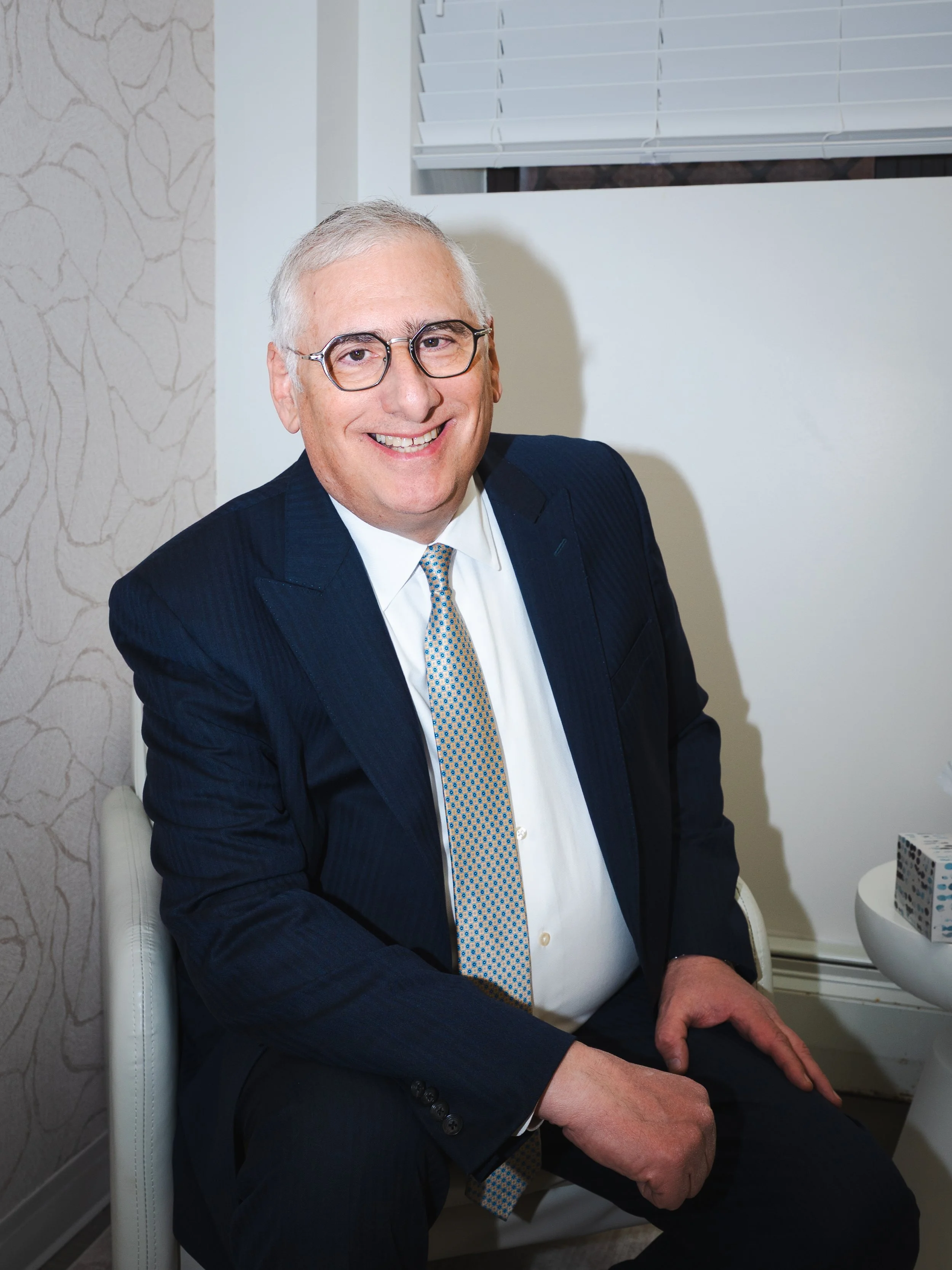 A smiling man with gray hair and glasses, wearing a dark suit, white shirt, and patterned tie, sitting indoors near a window with white blinds.
