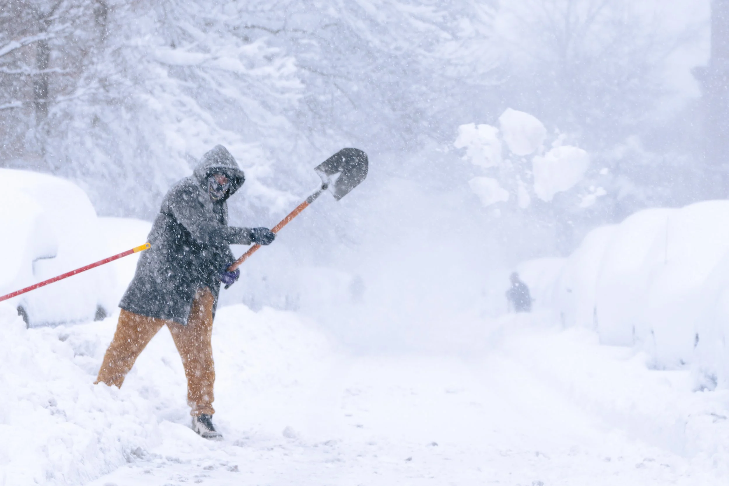 Person shoveling snow in a snowstorm, wearing a gray hooded jacket, tan pants, and gloves.