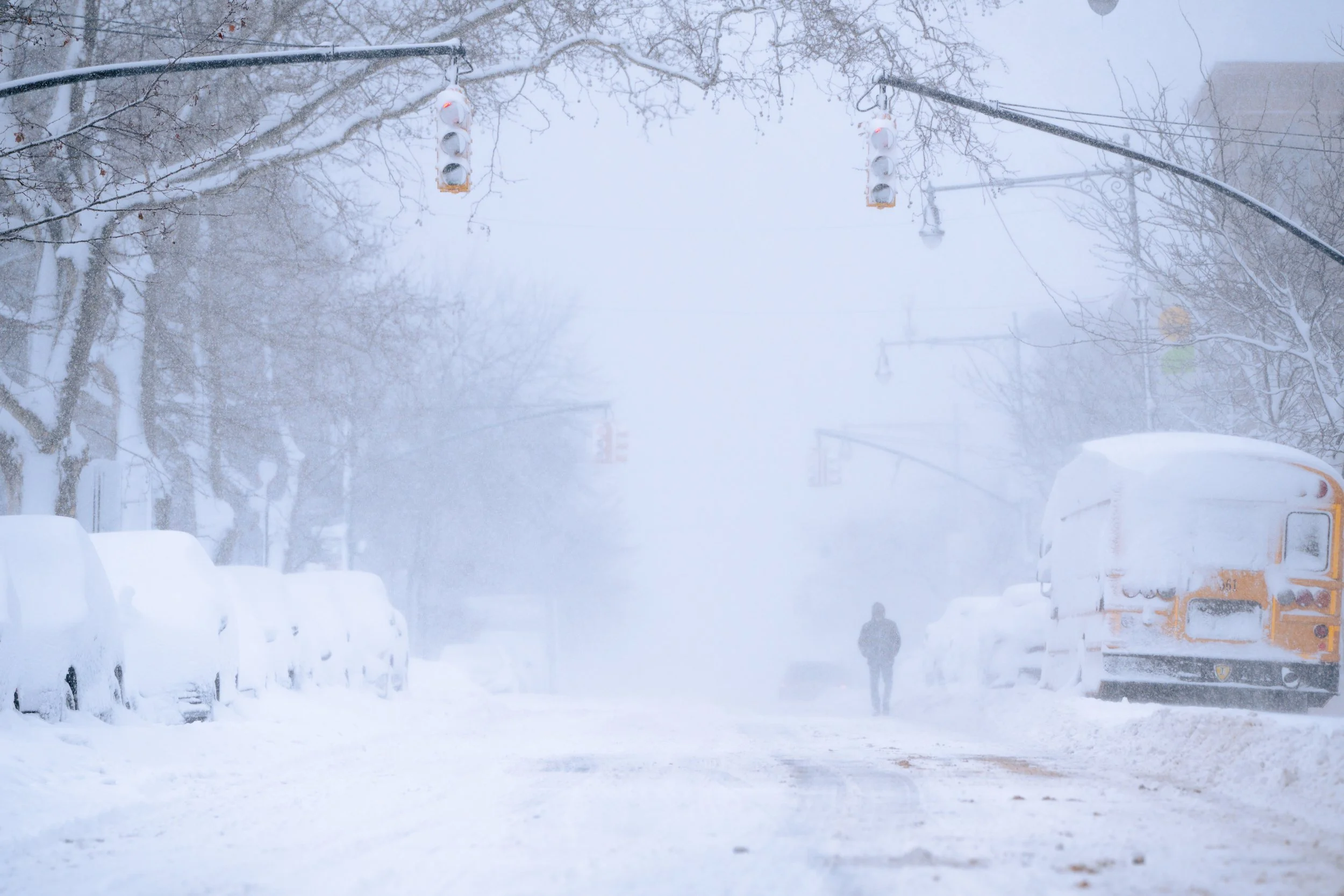 A snowy street scene with snow-covered cars parked along the sides, snow falling heavily, a person walking in the distance, and traffic lights hanging above the street amid foggy weather.