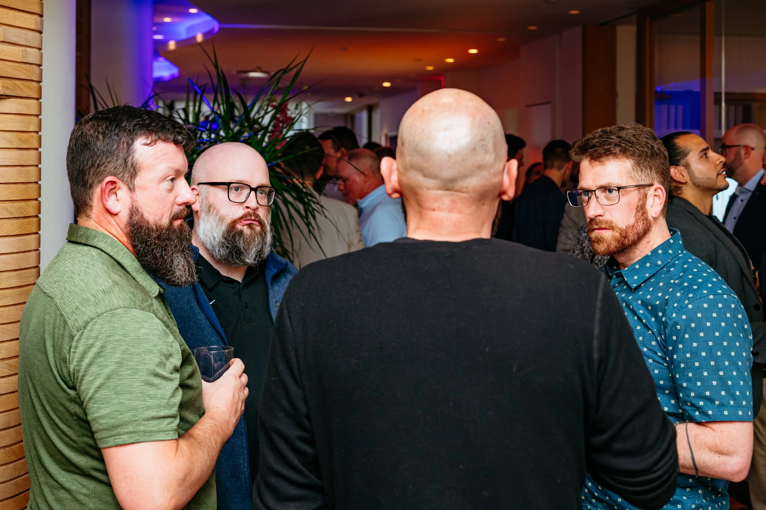 Group of men socializing at an indoor event, some holding glasses, in a room with warm lighting and wooden decor.