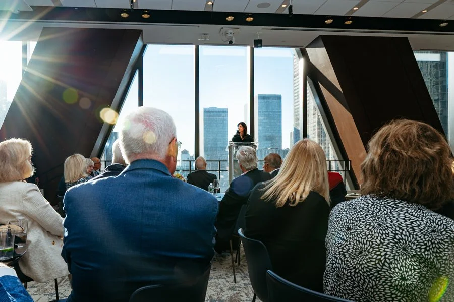 Business conference or presentation in a high-rise office with an audience of professionals, a presenter at a podium, and city skyline through large windows