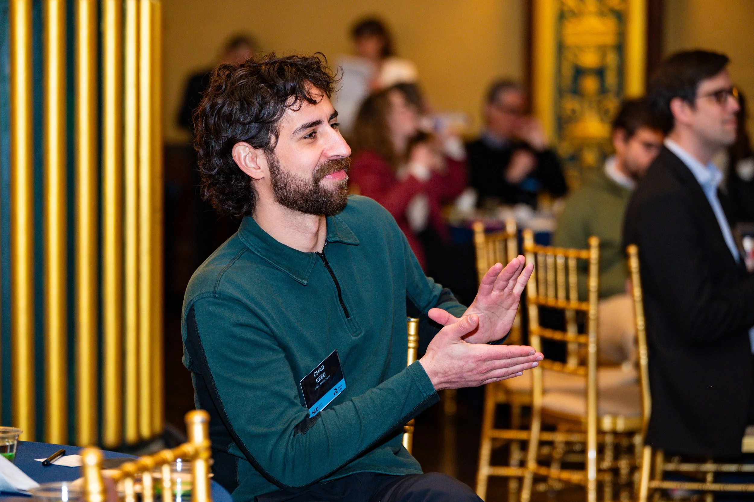 A man with a beard and curly hair, wearing a green zip-up jacket, is seated at a conference or event, clapping his hands. He has a name tag that reads 'Chad Reed.' There are other people seated at tables in the background.