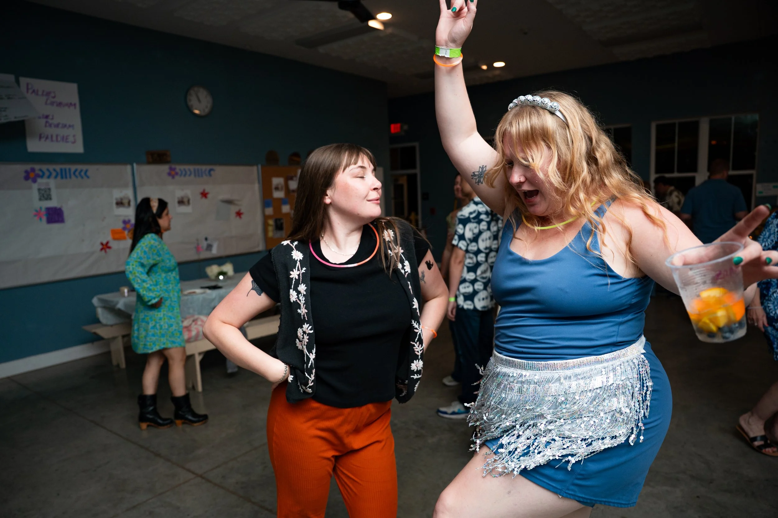 Two women dancing and having fun at a party or celebration in an indoor venue. One woman wears a blue dress with a silver, fringed skirt and a beaded headband, while the other is dressed in a black top with orange pants. Other people and posters are visible in the background.