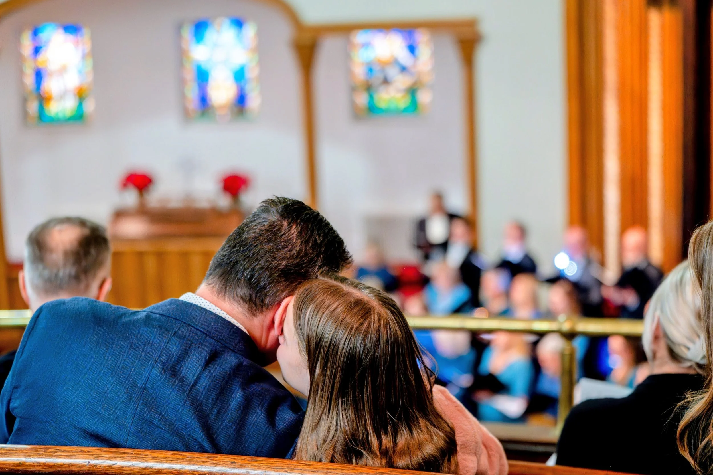 A couple sitting close together in a church pew, whispering or sharing a private moment during a formal event. The church interior features stained glass windows and people dressed in formal attire seated in the background.