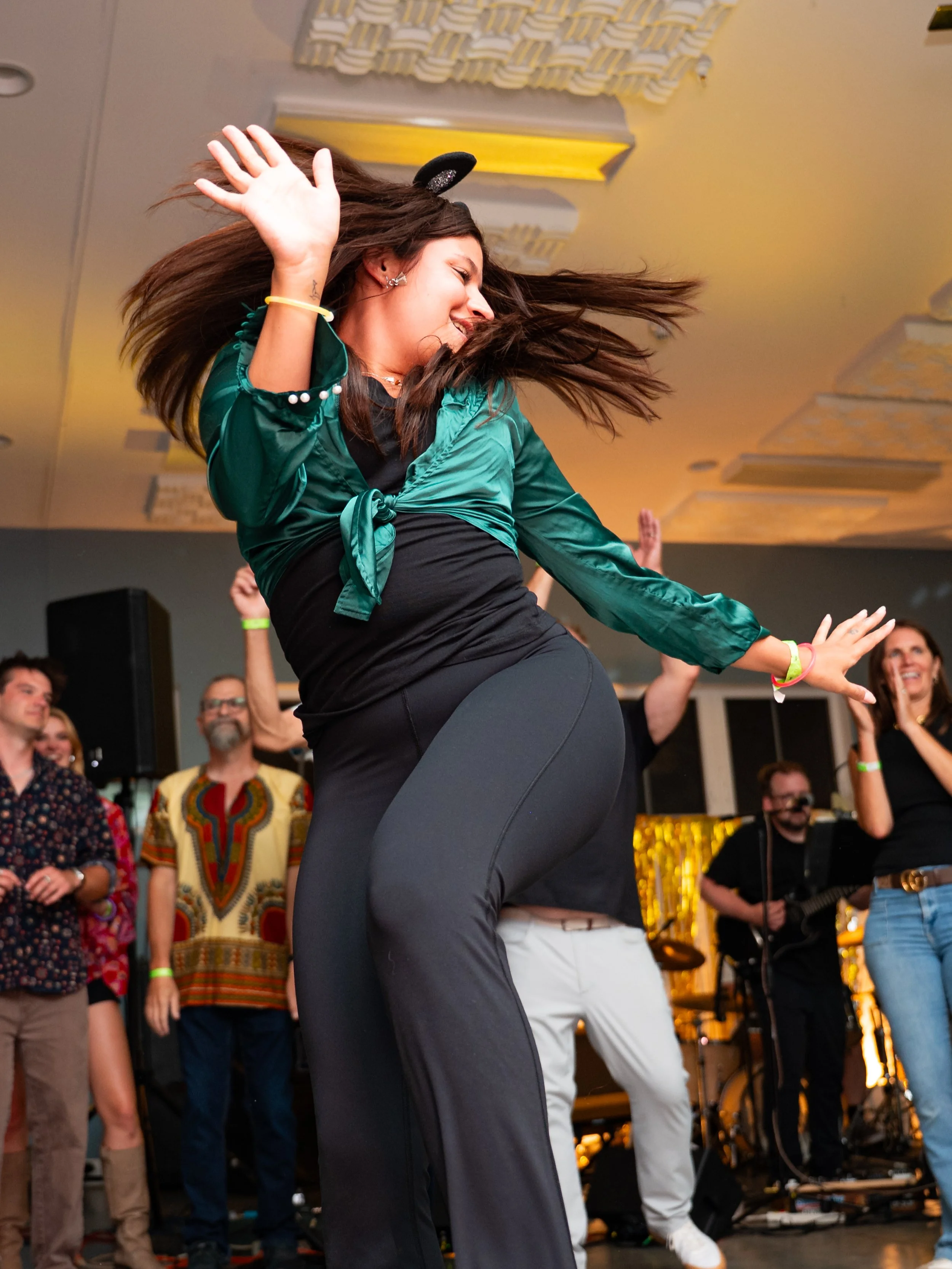 A woman dancing with her hair swinging, wearing a green satin top and black leggings, at an indoor social event with other people watching and a band playing in the background.