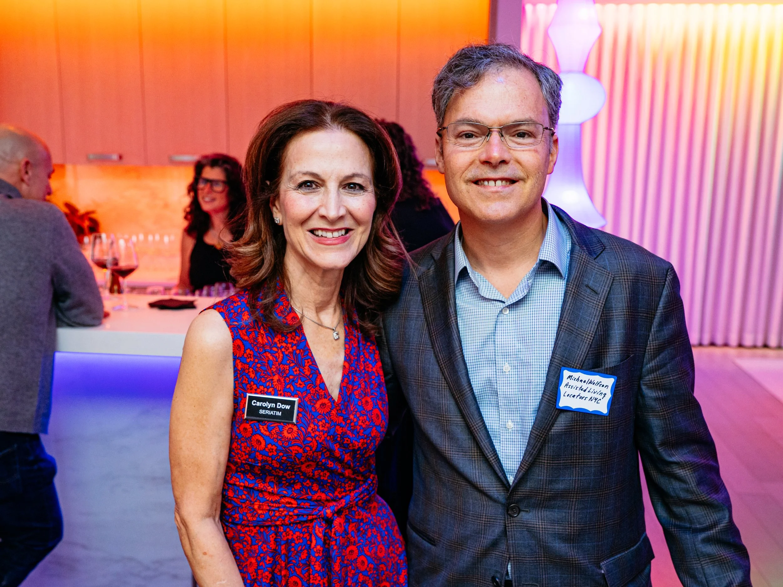 A woman and a man smiling at an indoor event. The woman has shoulder-length brown hair, is wearing a red and blue floral dress, and has a name tag that reads 'Carolyn Dow.' The man has short gray hair, glasses, and is wearing a gray checkered blazer 