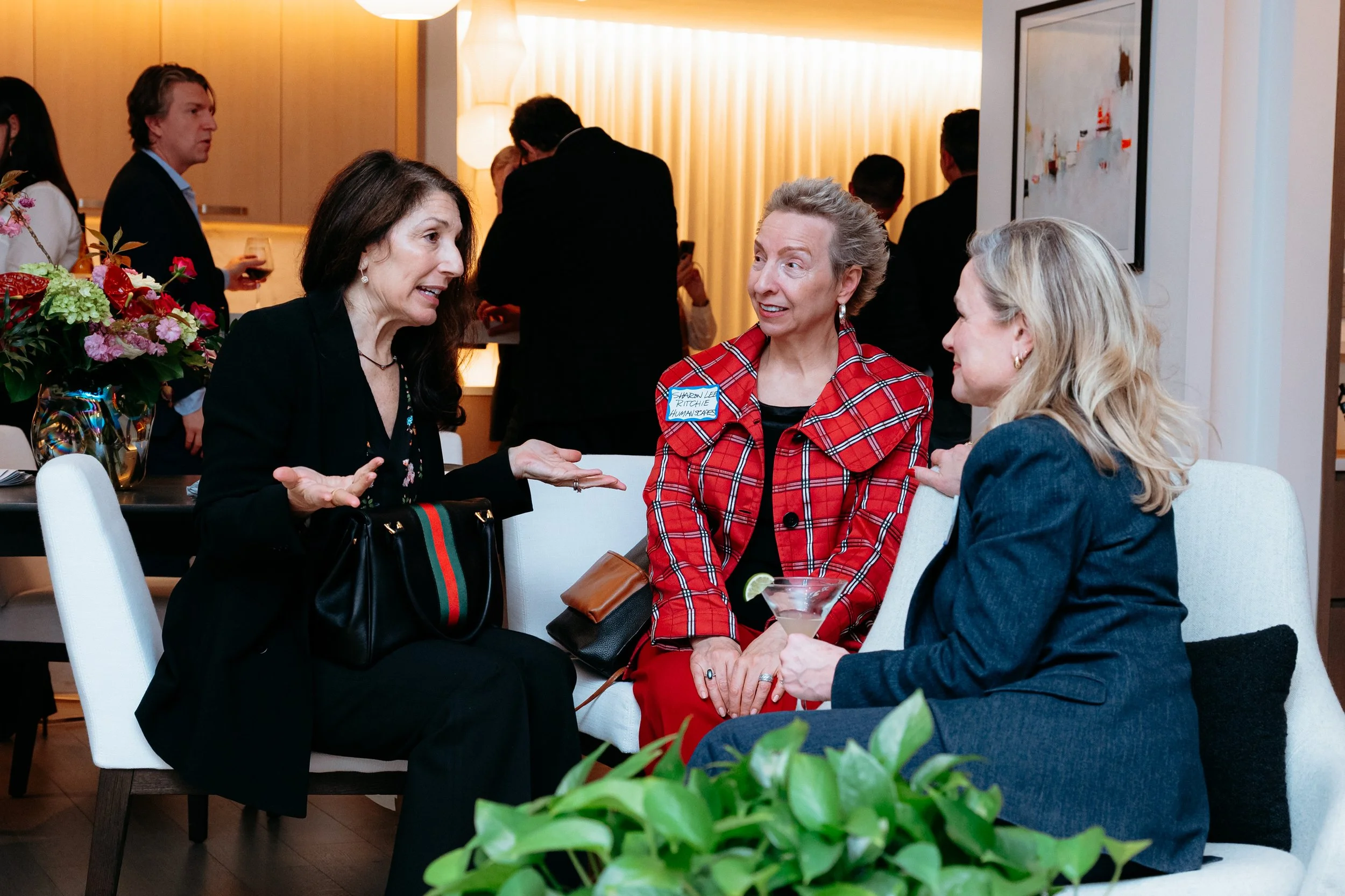 Three women are engaged in a conversation at a social event indoors. One woman is speaking animatedly, while the other two listen attentively. There are drinks and floral arrangements on the table. In the background, other people are mingling and hol