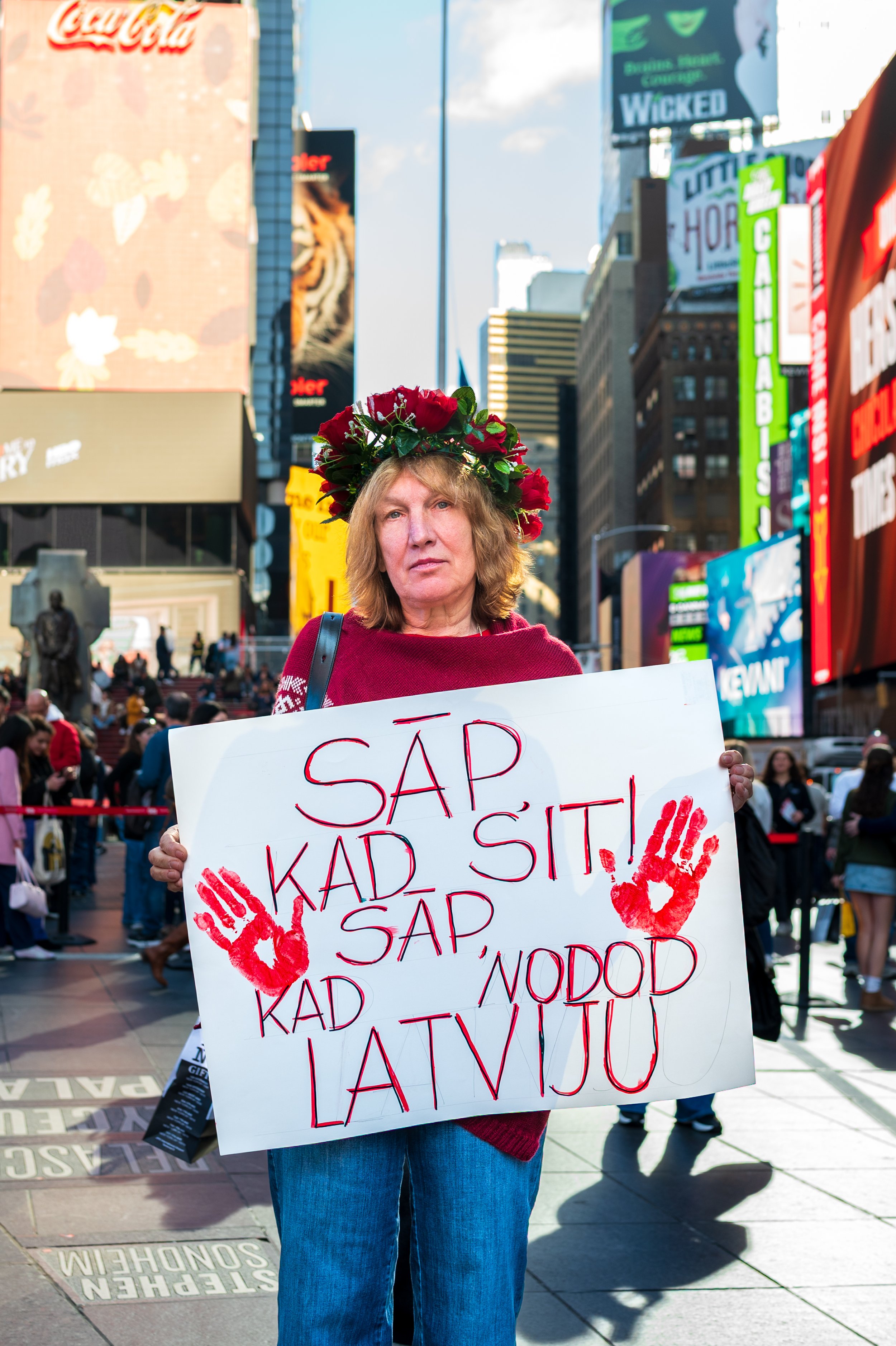 A woman standing in Times Square holding a protest sign. She wears a floral crown and a red sweater, with a serious expression. The sign has red handprints and text in Latvian.
