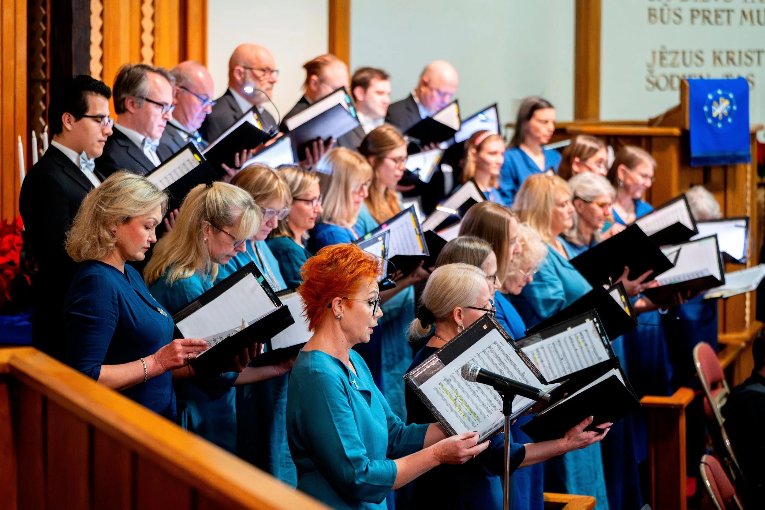 A choir of men and women singing in a church or auditorium, holding sheet music, with some wearing blue attire and others in black suits, and a microphone stand in front.