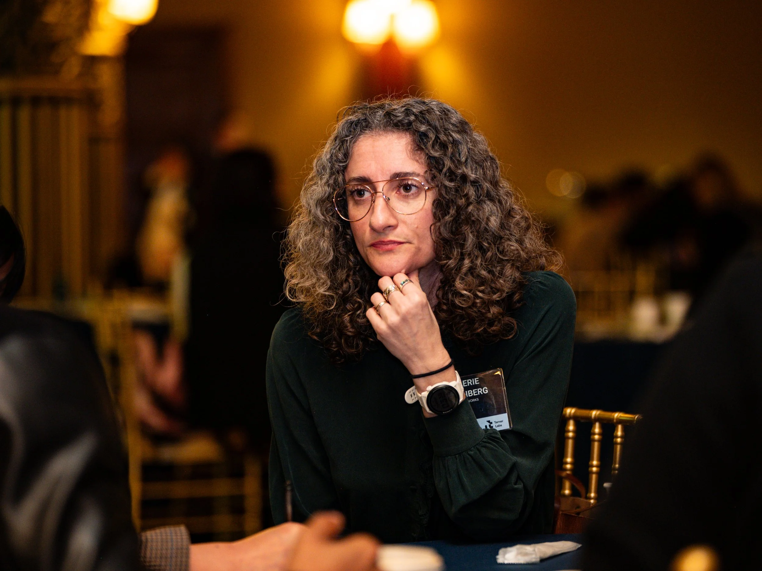 A woman with curly hair and glasses sitting at a table in a dimly lit room, wearing a black top and a watch, with her hand resting on her chin, appearing to be deep in thought.