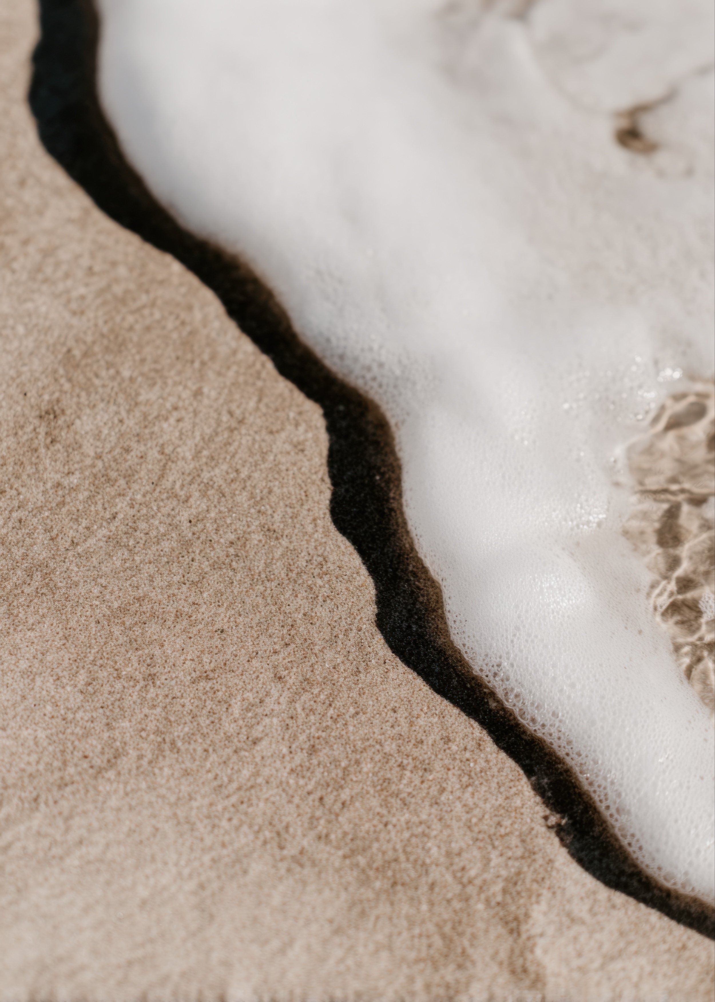 Close-up of sea foam washing onto sandy shoreline with warm natural tones and gentle water detail.