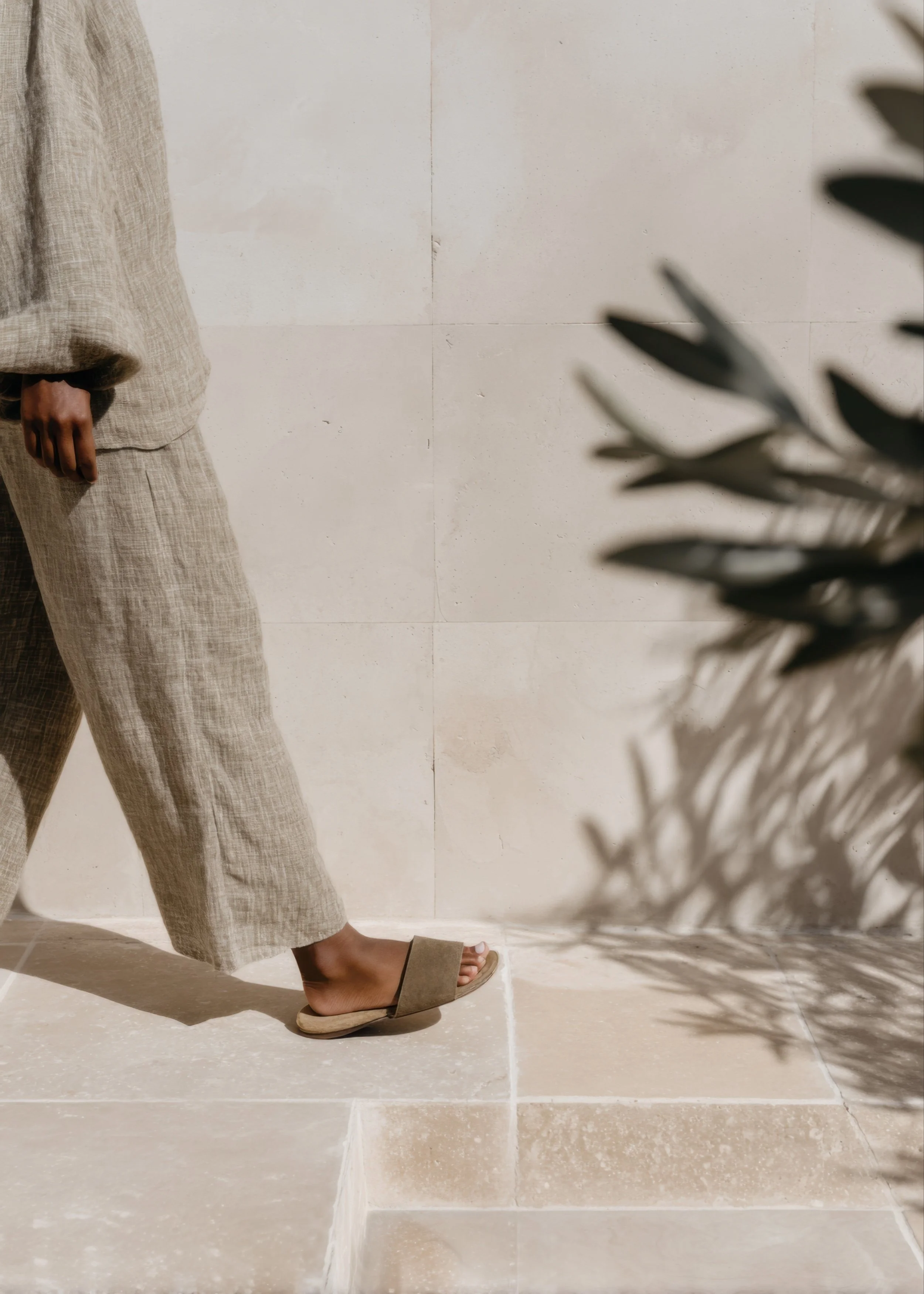 Person walking in neutral linen clothing and sandals beside a sunlit stone wall with soft plant shadows
