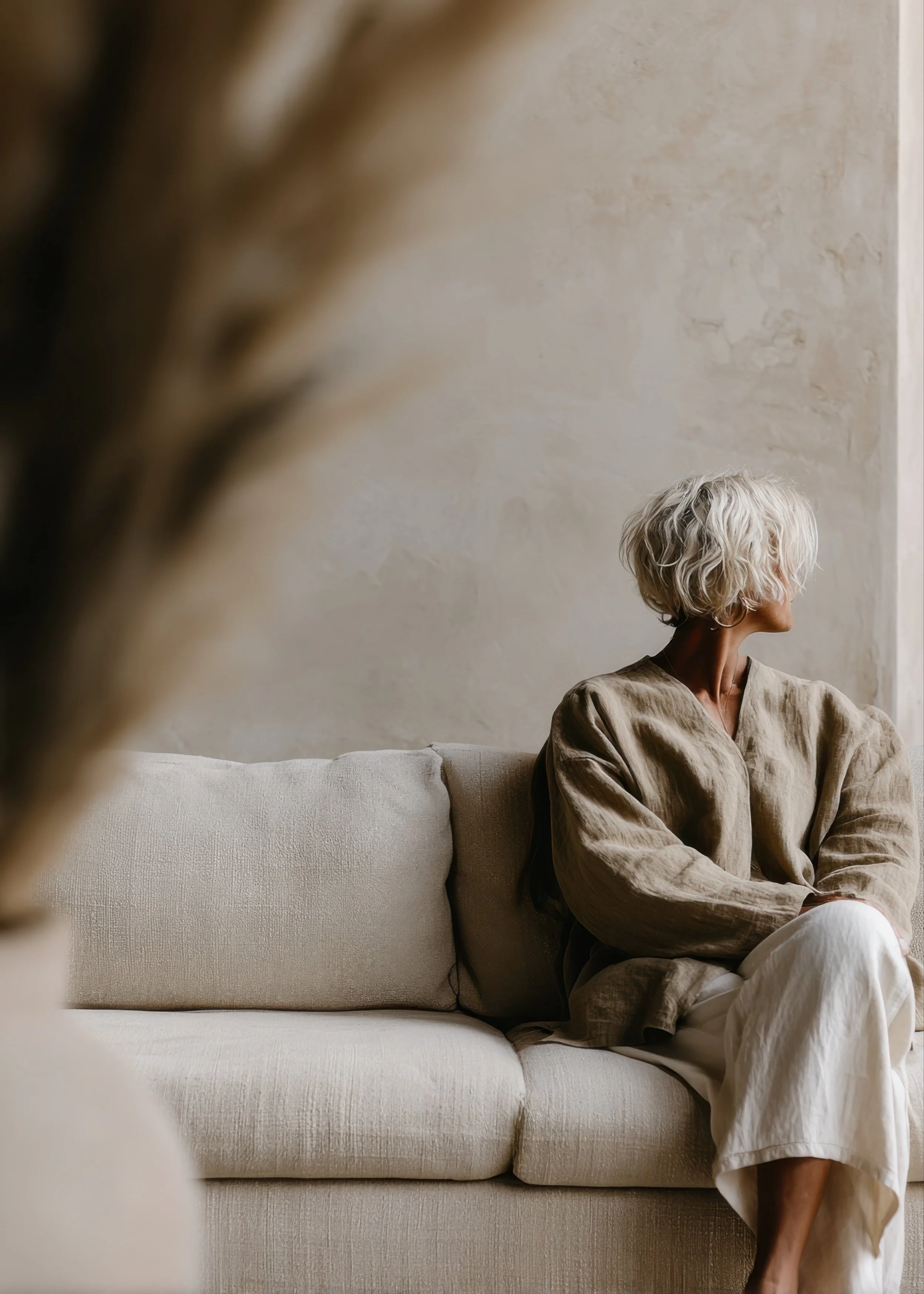 Person seated on a neutral sofa in a calm, sunlit room, looking toward the window with a reflective expression