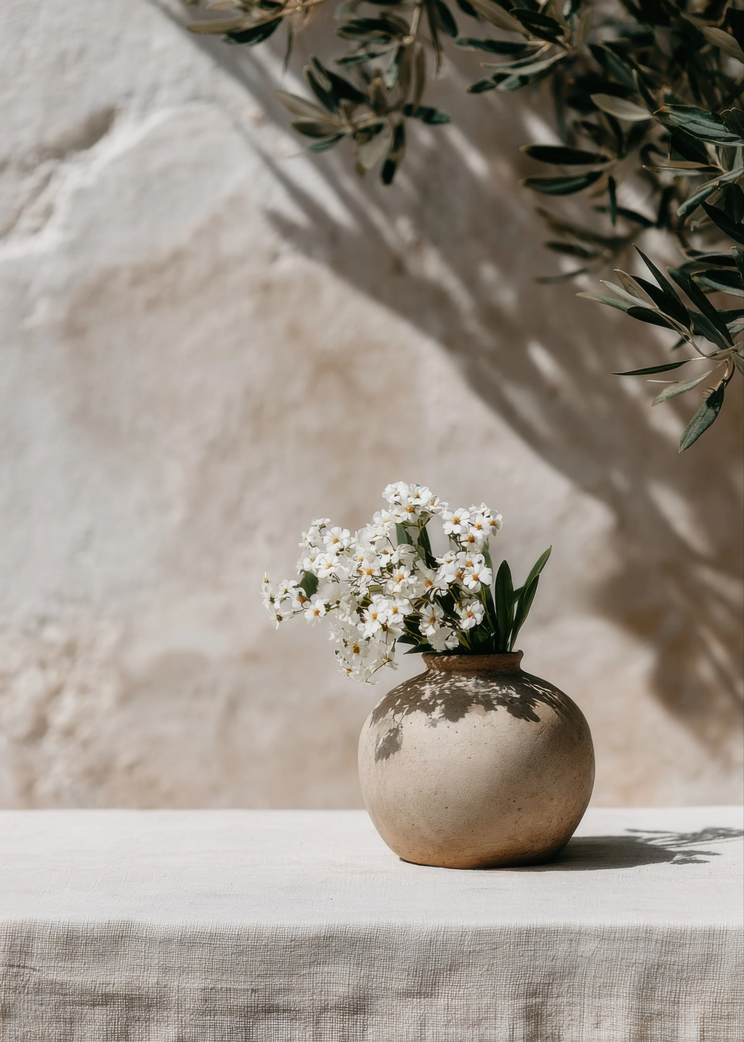 Ceramic vase with white flowers on a linen-covered table against a sunlit neutral wall with soft olive branch shadows