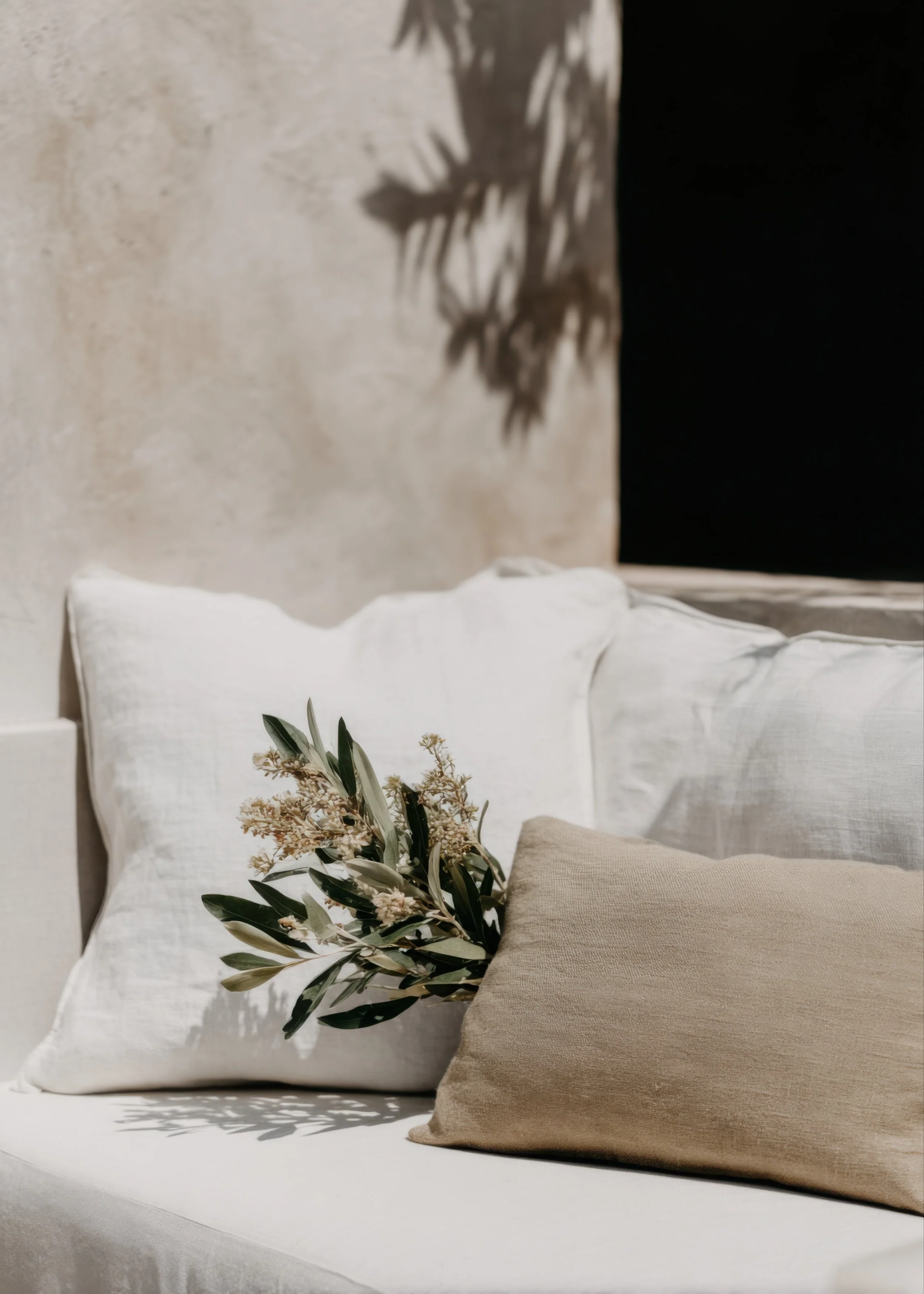 Neutral-toned pillows and greenery arranged on a light outdoor sofa with soft sunlight and plant shadows in the background