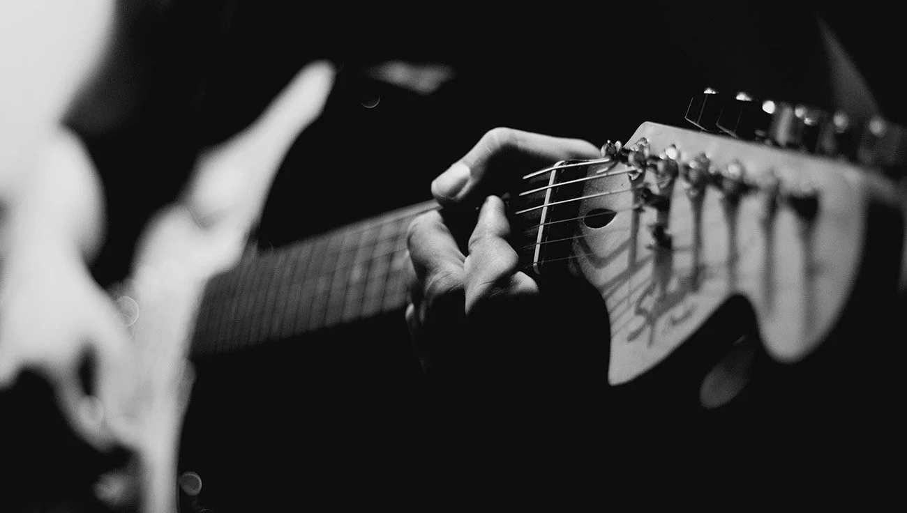 Close-up black and white photo of a person playing an electric guitar, focusing on the hand and guitar strings.