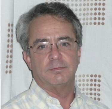 Man with glasses and gray hair wearing a plaid shirt in front of a patterned curtain.