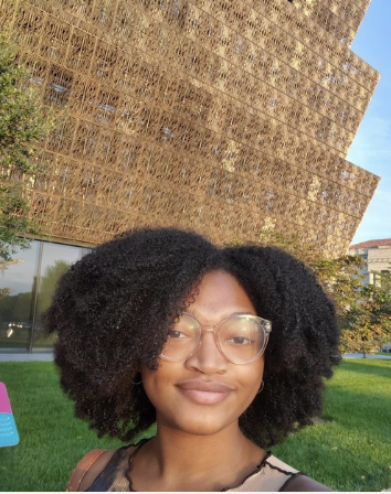 Person with curly hair and glasses in front of a modern architectural building with a patterned exterior.