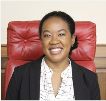 Person sitting on a red chair, smiling, wearing a black blazer and white shirt.