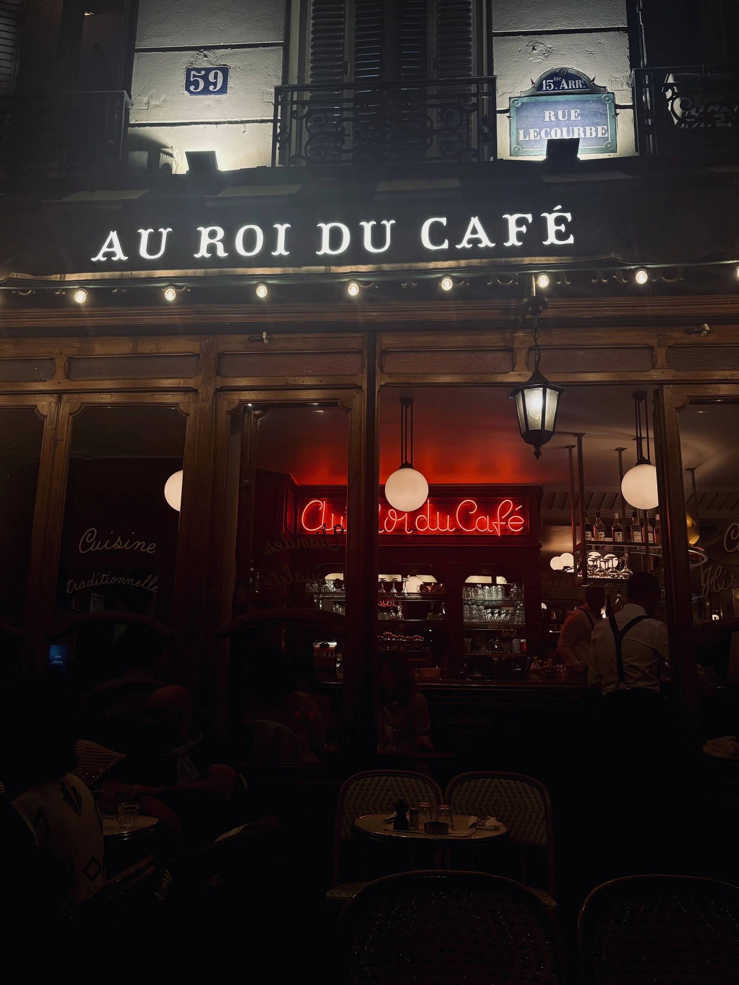 Nighttime view of Au Roi du Café, with a bright white sign, red neon, warm wooden windows, and patrons inside; the street café feels cozy and lively.