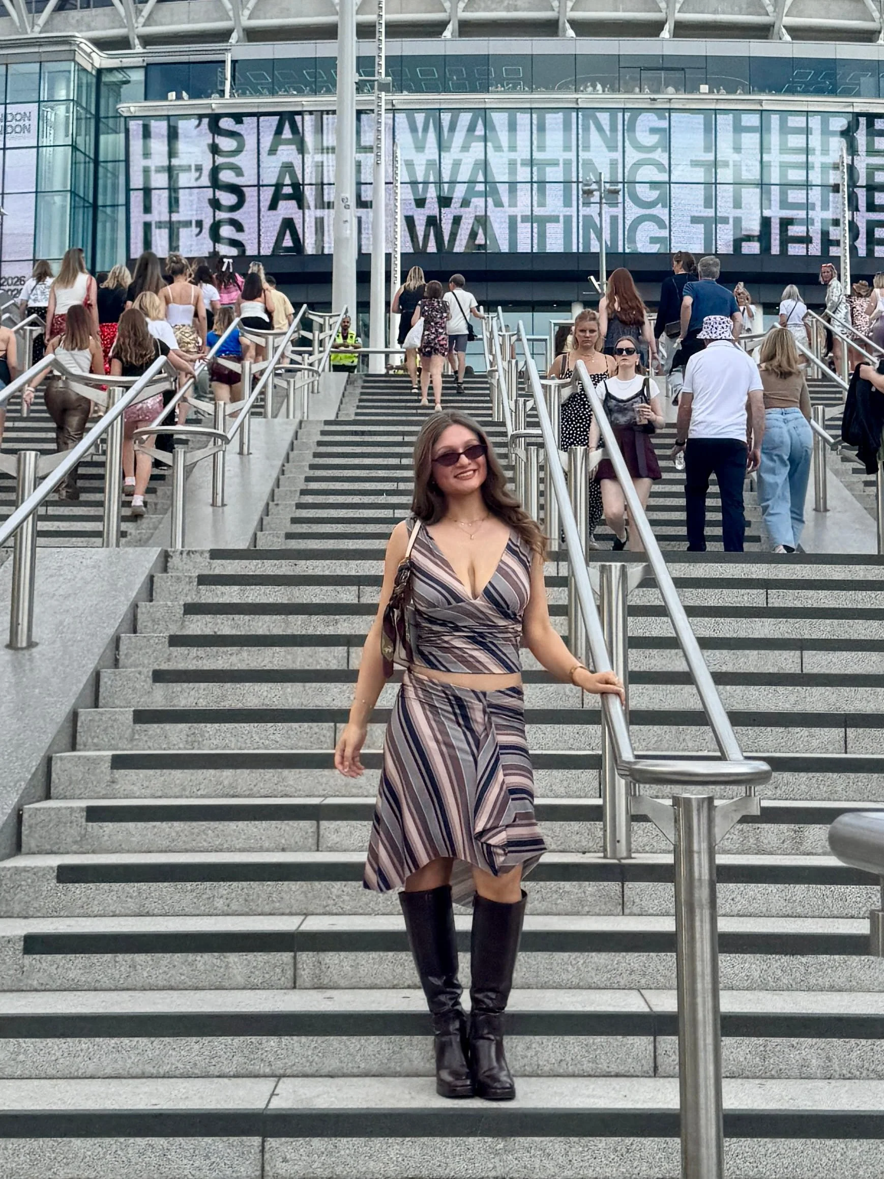 A woman wearing a striped two-piece and boots poses on stairs, in front of Wembley stadium.