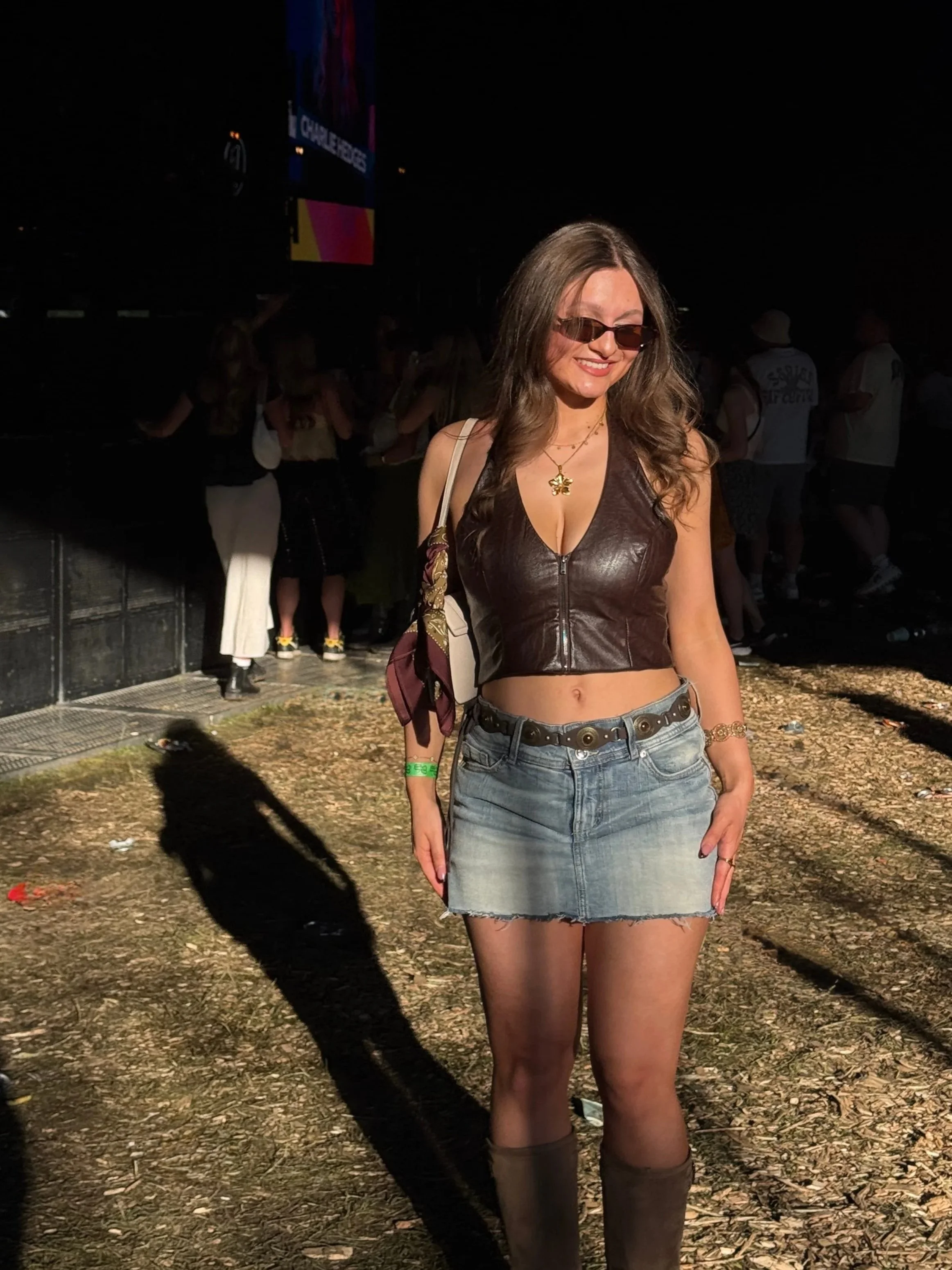 Woman wearing a brown leather crop top, denim skirt, and boots standing in a sunlit outdoor event with people in the background.