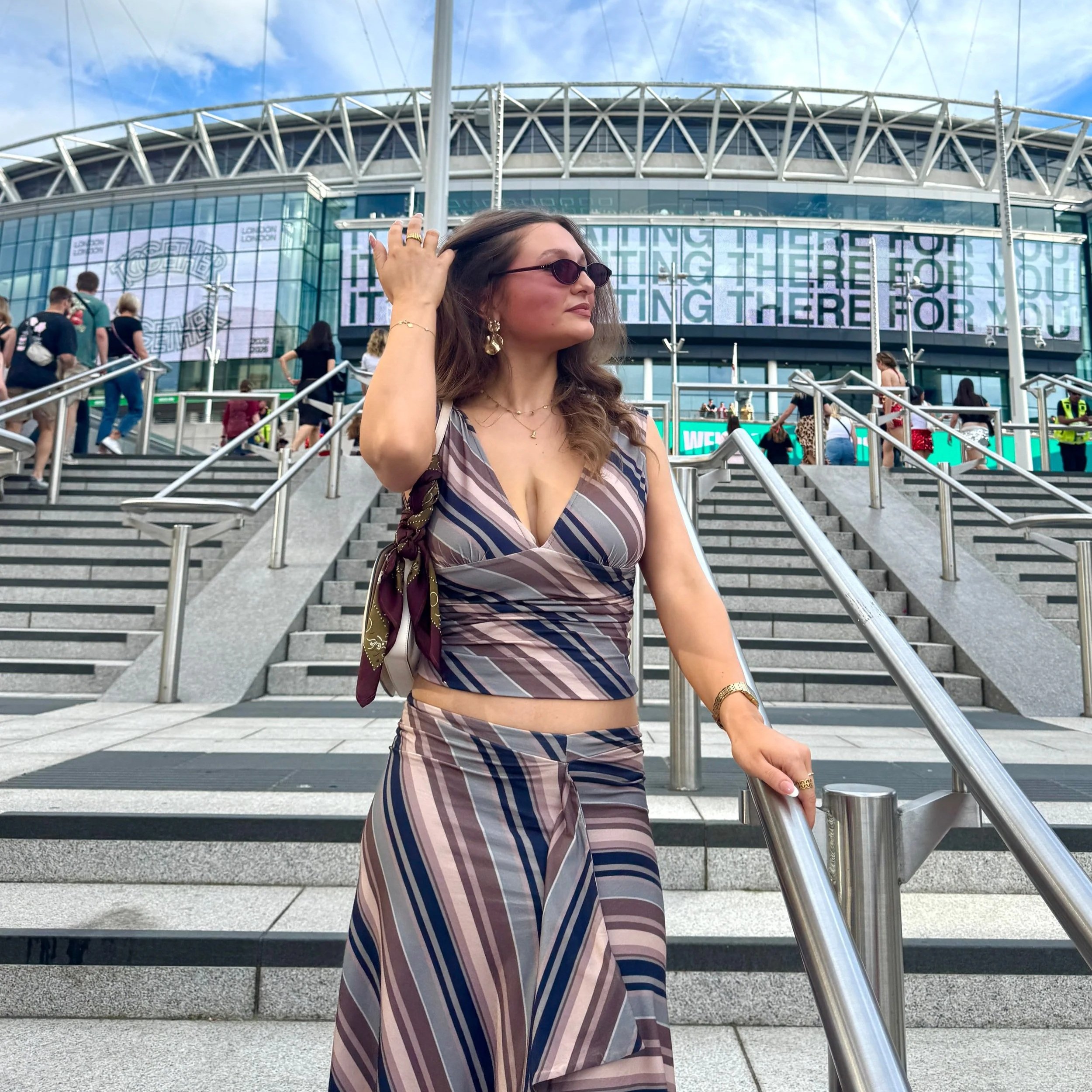 A woman in a striped top and sunglasses descends stairs, enjoying the vibrant atmosphere of a Harry Styles concert at Wembley.