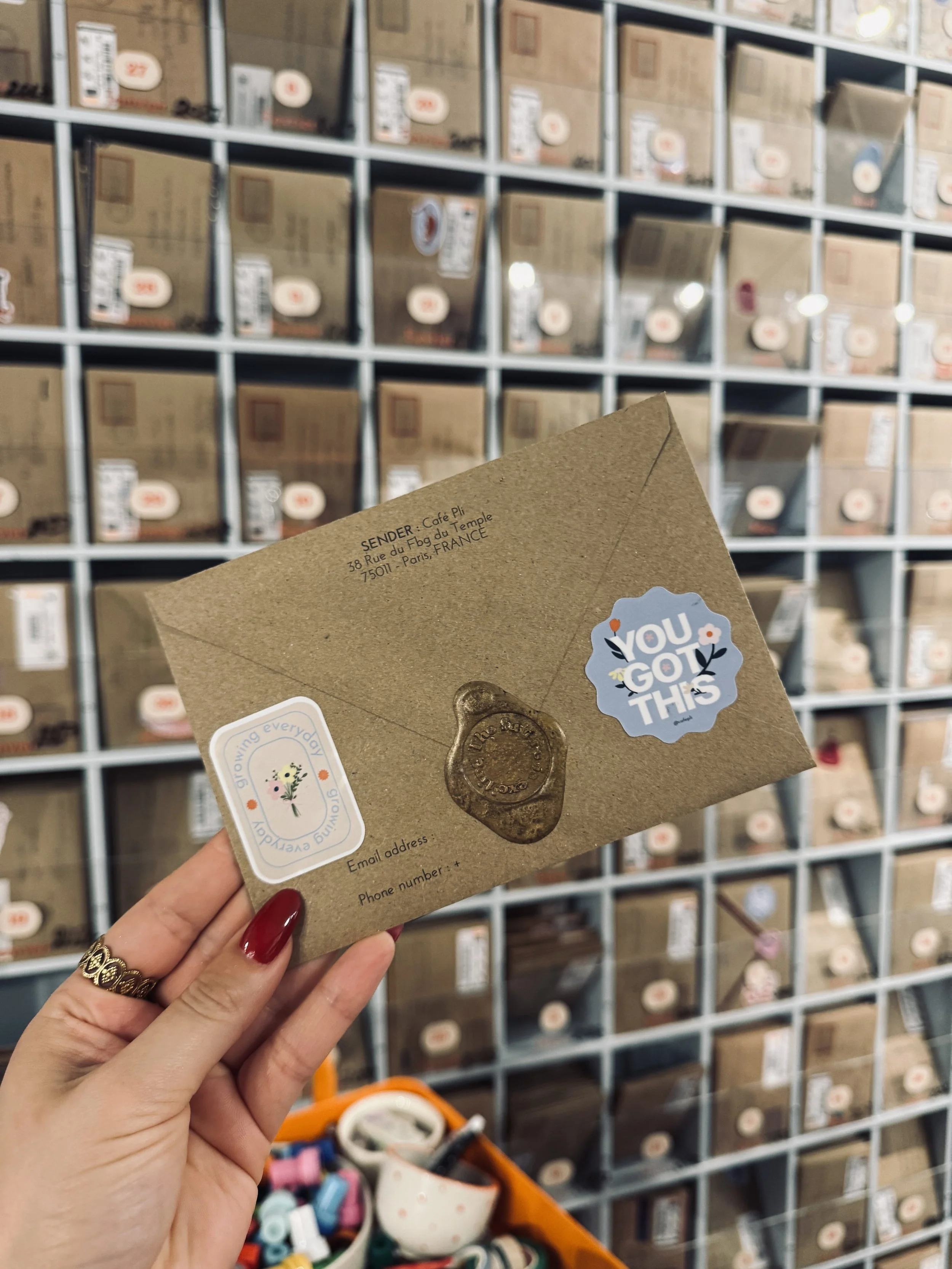 A hand with red nails holds a kraft envelope with a wax seal and “You got this” sticker before rows of neatly boxed goods in a cozy cafe..