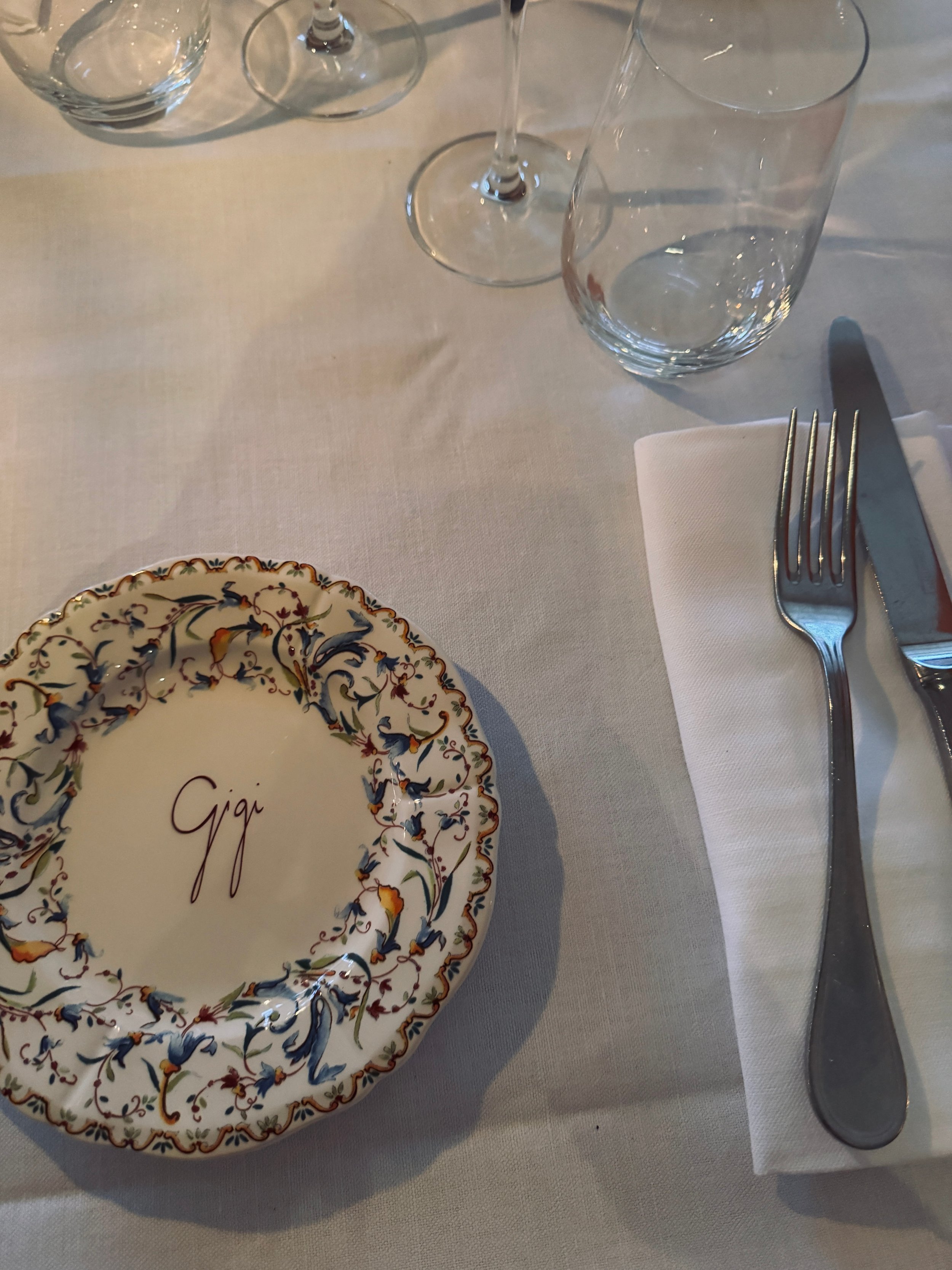 An elegant restaurant place setting features a floral plate marked “Gigi,” clear glasses, and polished cutlery on a white linen tablecloth.