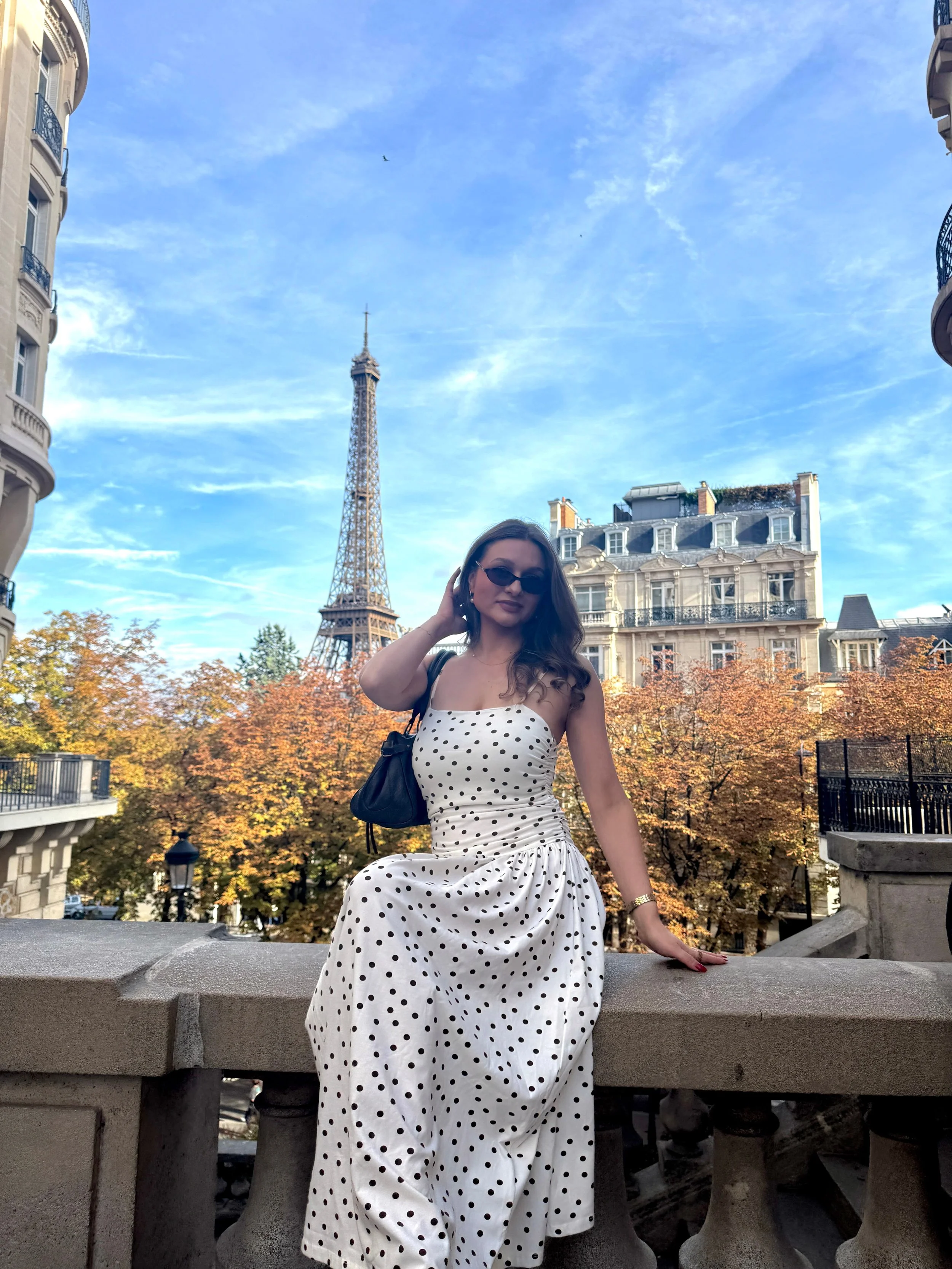 Woman in a white polka-dot dress poses on a Paris balcony with the Eiffel Tower behind her, framed by autumn trees under a blue sky.