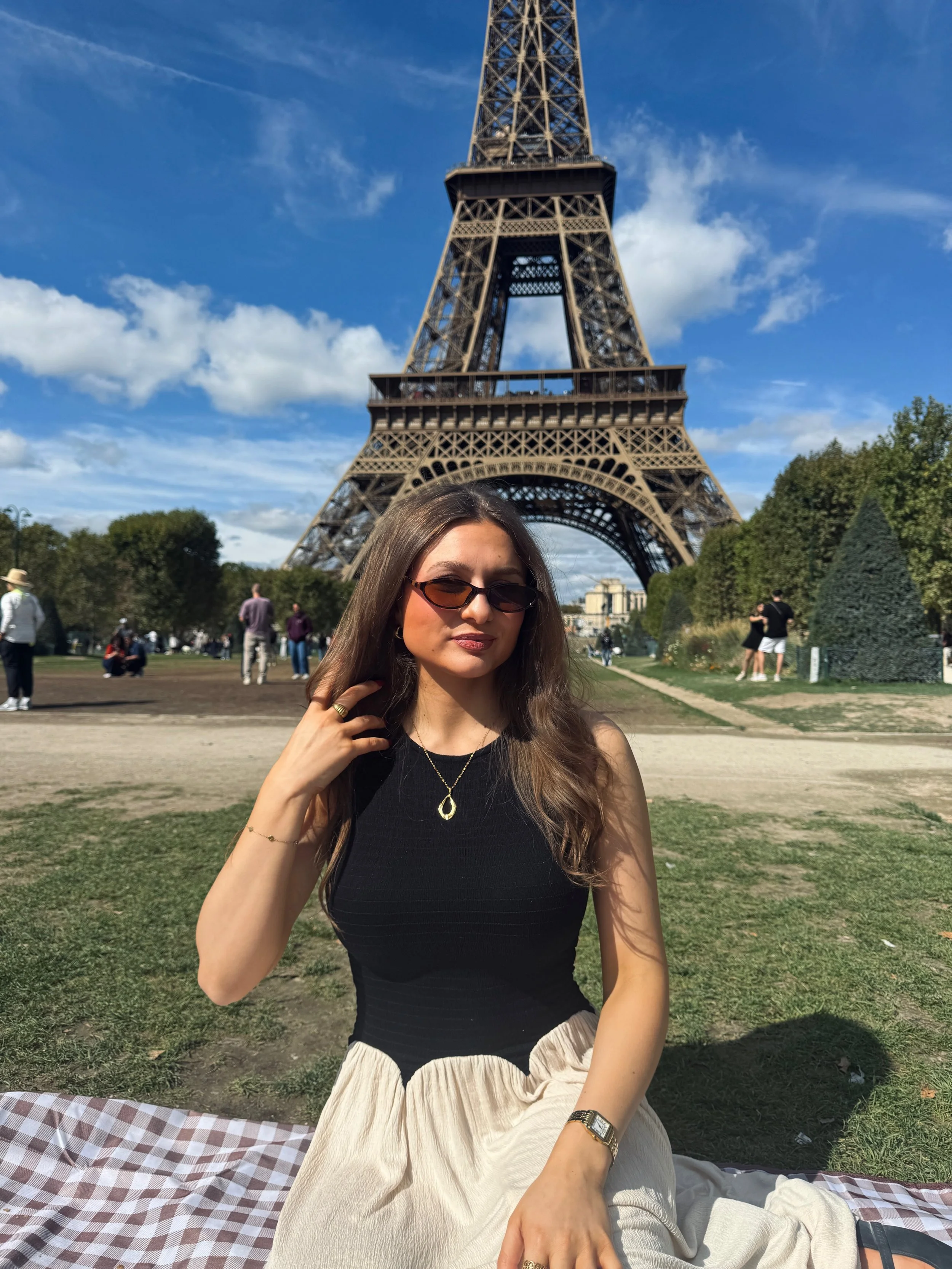 Woman in sunglasses sits on a picnic blanket before the Eiffel Tower in Paris, smiling on a sunny day with blue skies and park greenery.
