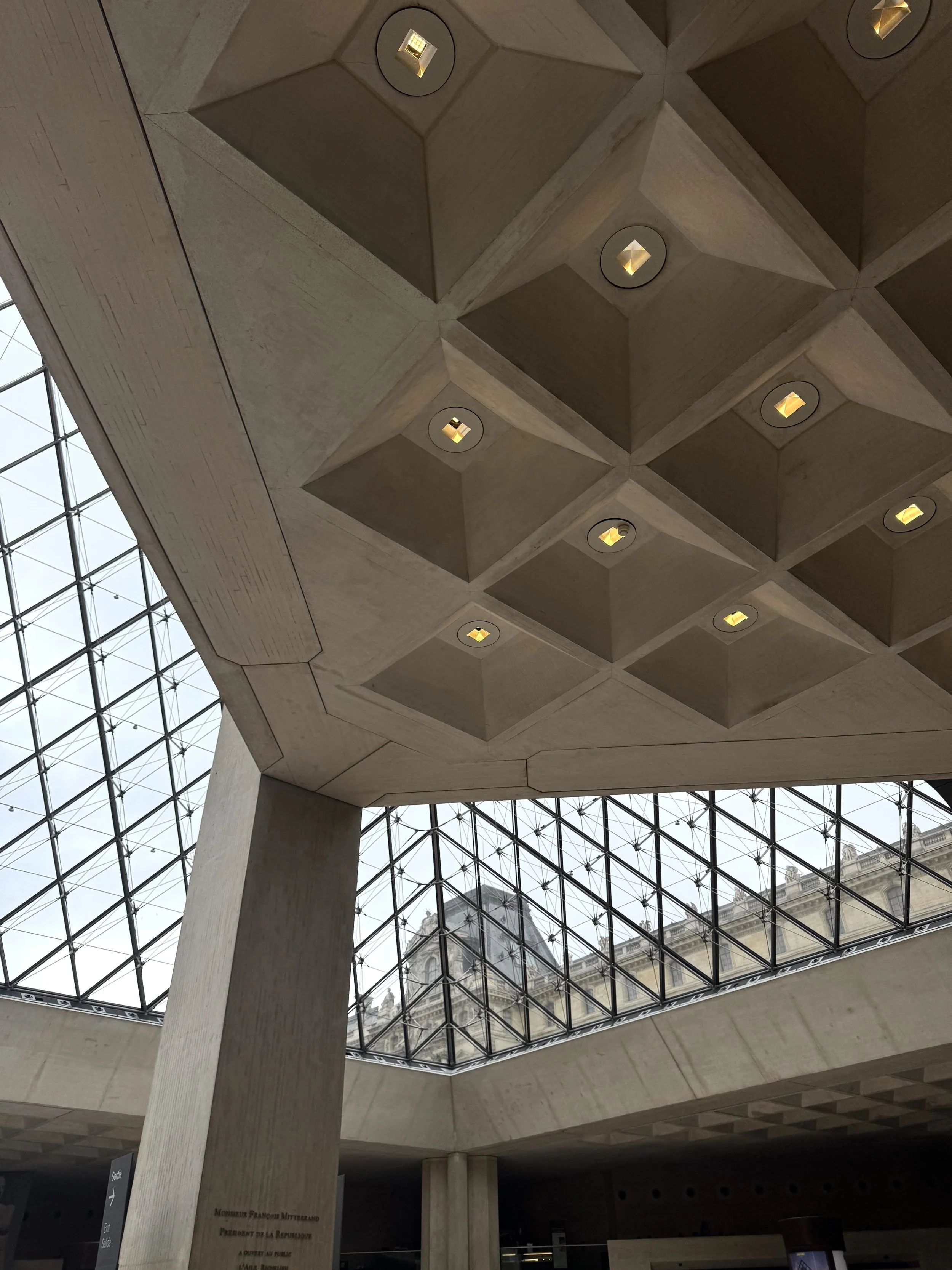 A monumental concrete interior with a geometric coffered ceiling and small amber lights, framed by a vast glass-and-steel skylight; quiet, austere atmosphere.