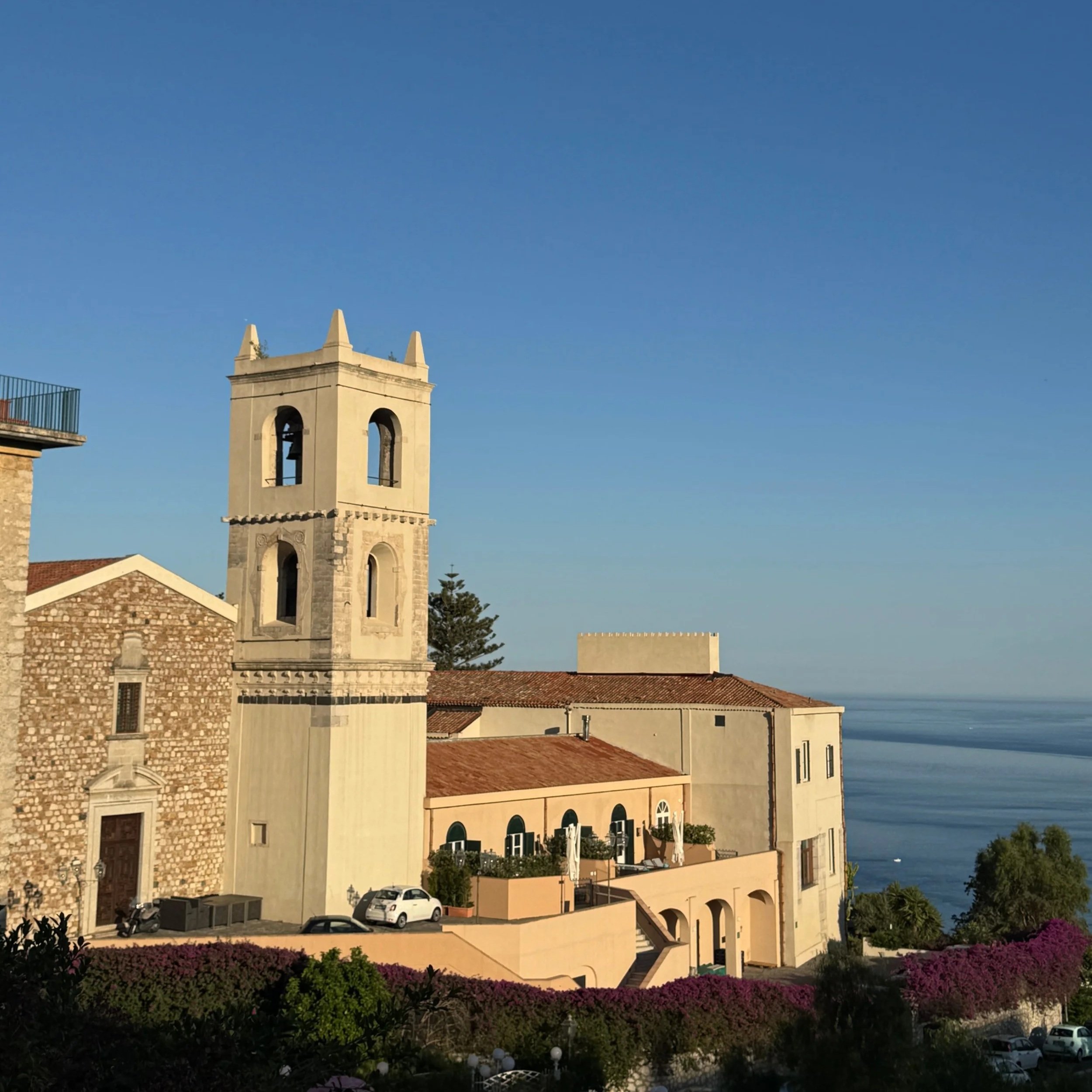 Sunlit Mediterranean buildings with a bell tower overlooking the sea under a clear blue sky.