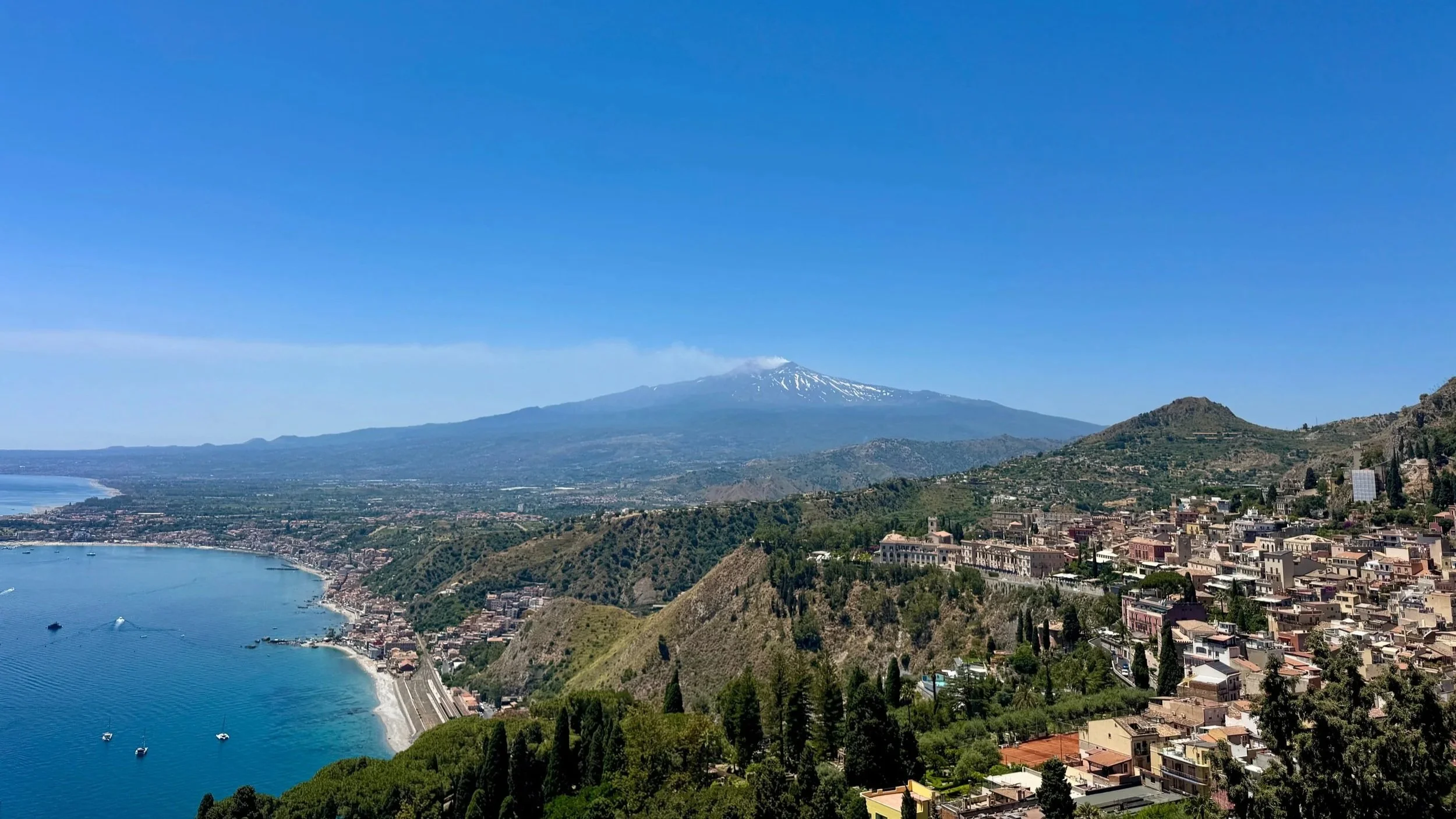Panoramic coastal town view with blue sea, lush hills, and snow-capped mountain under clear sky.