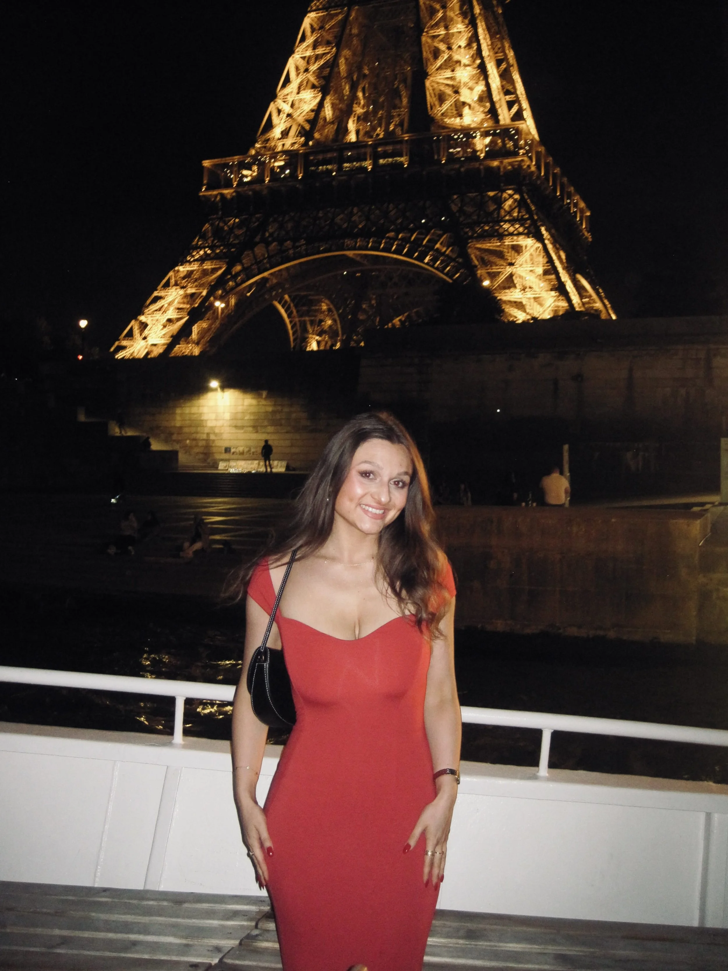 A smiling woman in a red dress poses on a riverside walkway at night, with the golden-lit Eiffel Tower rising behind her against a dark sky.