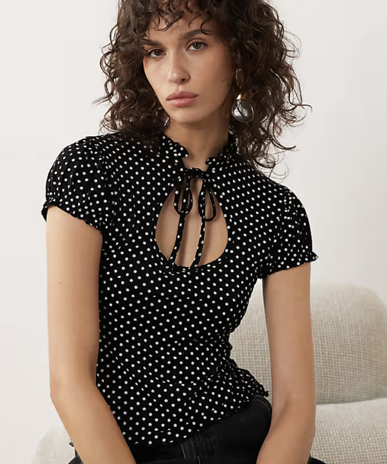 A woman with curly hair looks at the camera, wearing a black and white polka dot top with short sleeves and a keyhole neckline.