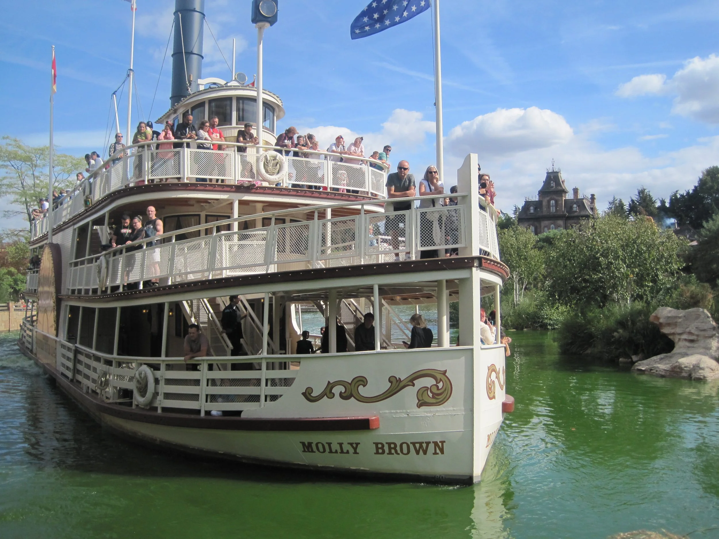 The Molly Brown riverboat, crowded with sightseeing passengers and flying flags, glides across green water in a sunny, landscaped park.