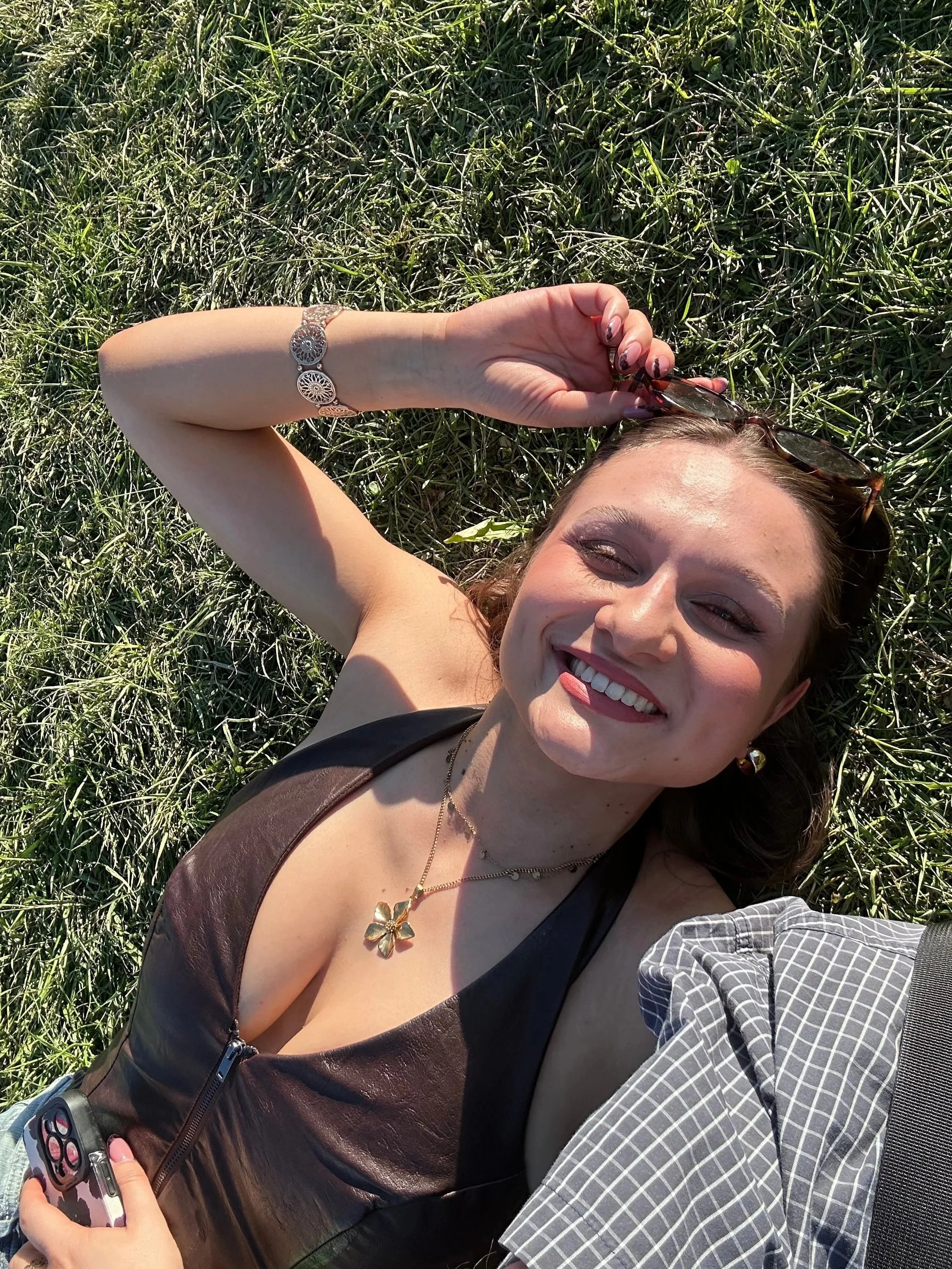 Woman lying on grass wearing a brown halter top, gold flower necklace, and bracelet, holding sunglasses and a phone.