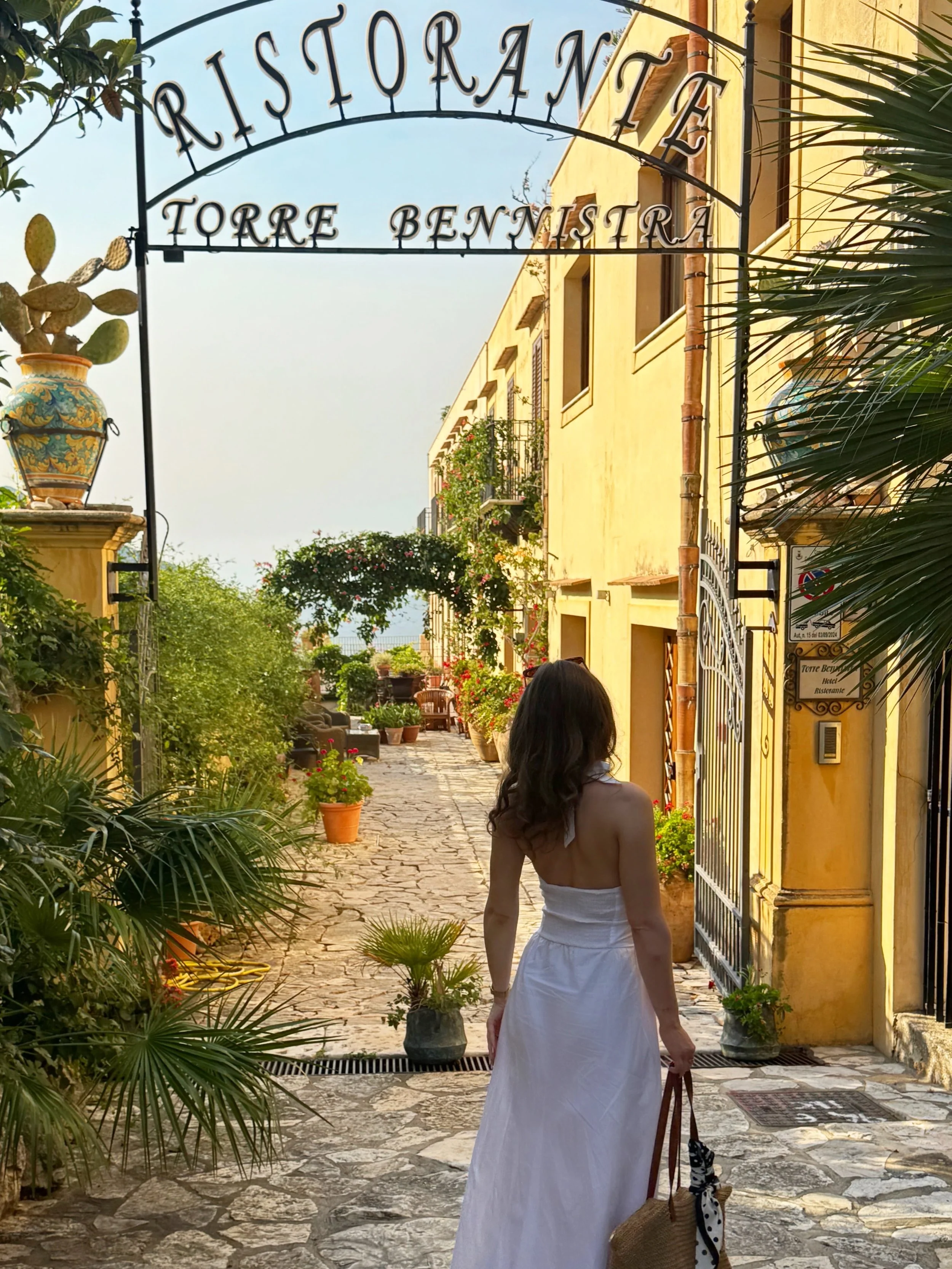 A woman in a flowing white dress strolls gracefully through a sunlit courtyard surrounded by lush greenery.