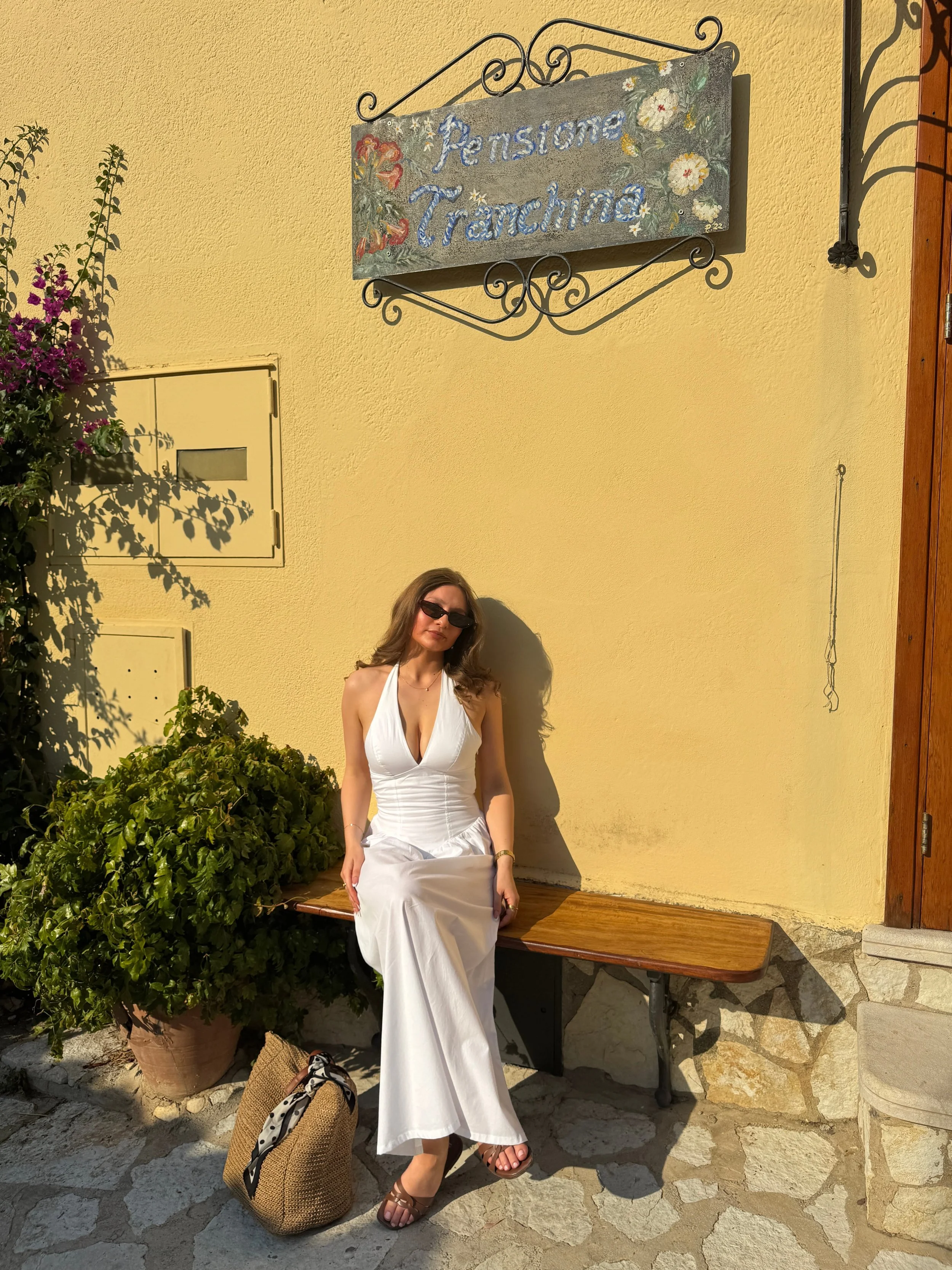 A woman wearing a white dress relaxes on a bench, surrounded by greenery, exuding a serene and tranquil vibe.