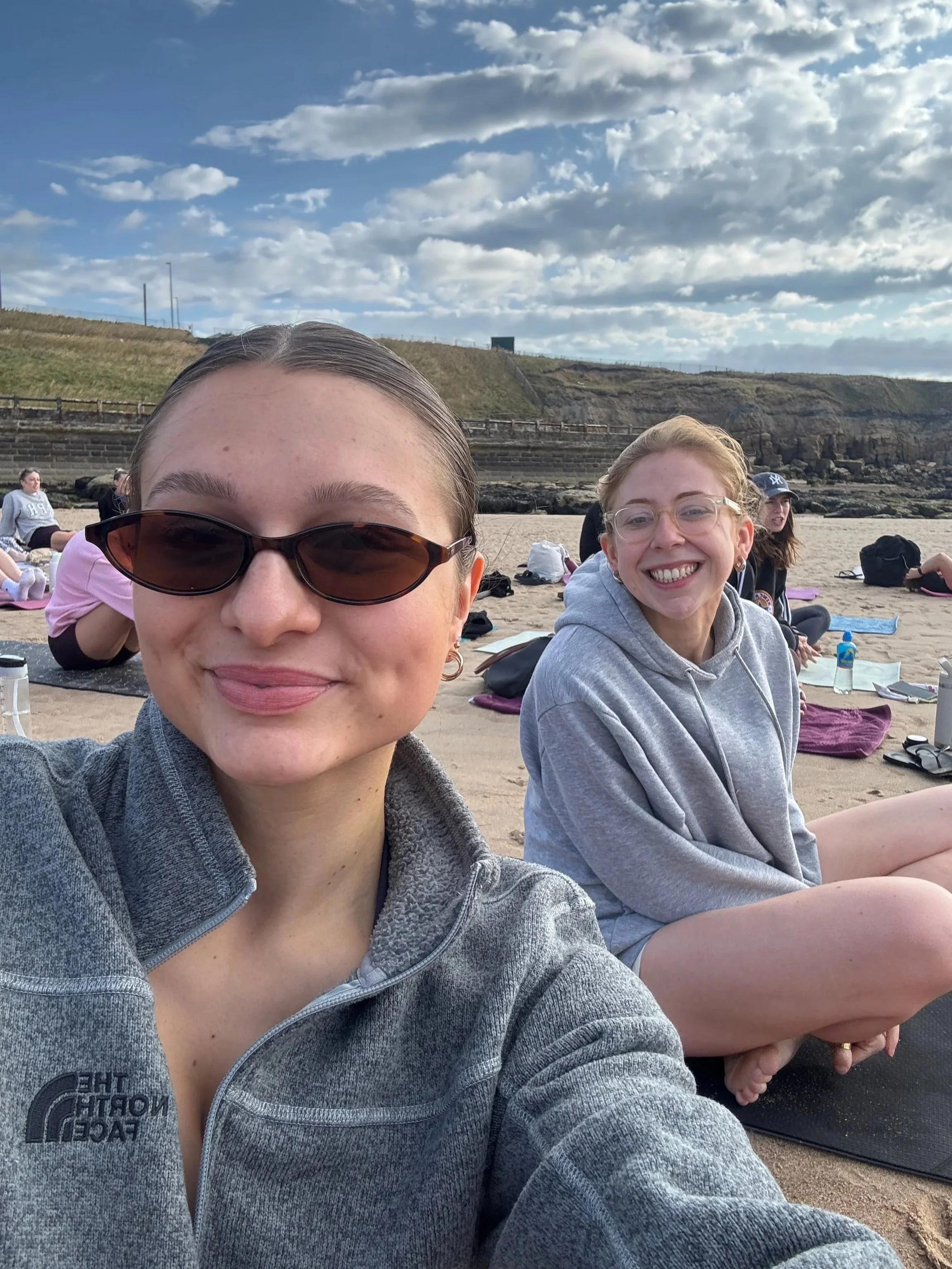 Two friends sit on the sandy beach, sporting stylish sunglasses and soaking up the sun.