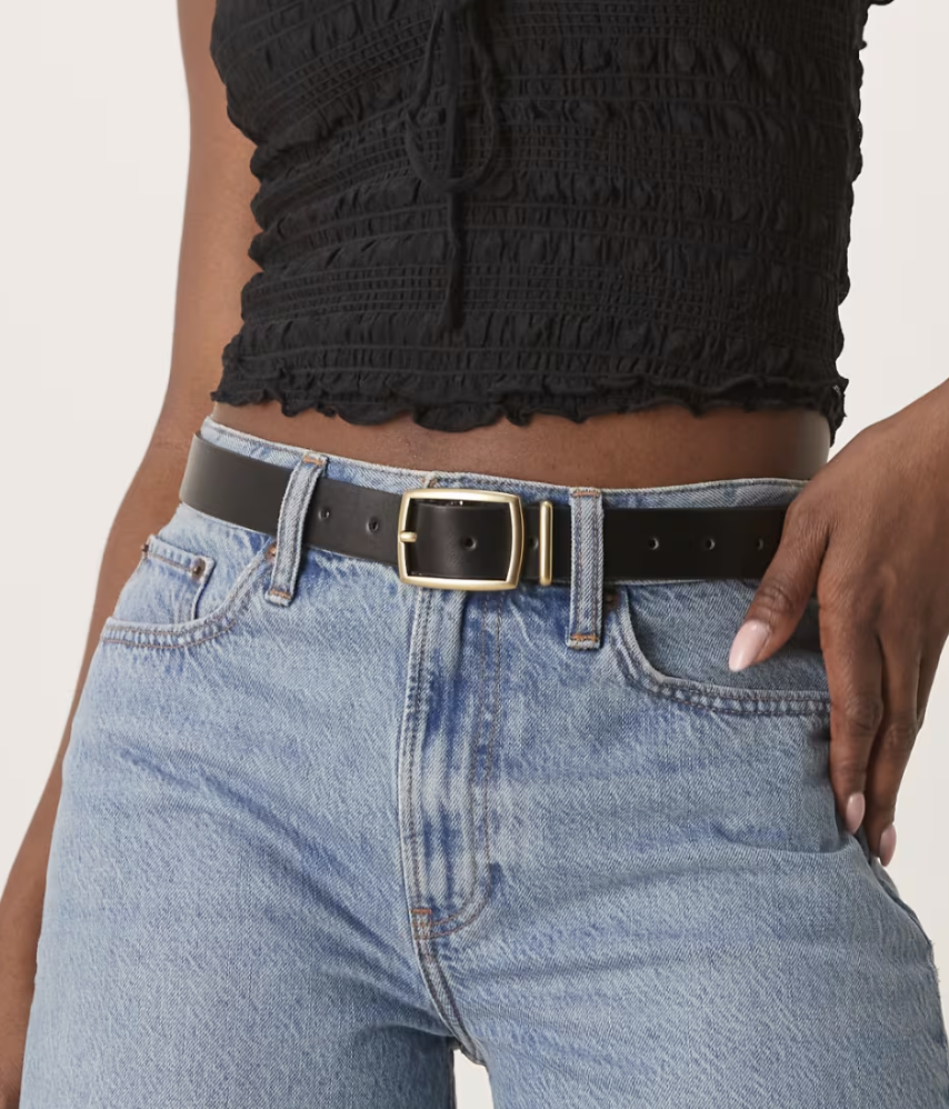 A person wearing a black smocked top and light blue jeans, with a black leather belt featuring a gold buckle. The style is casual and chic.
