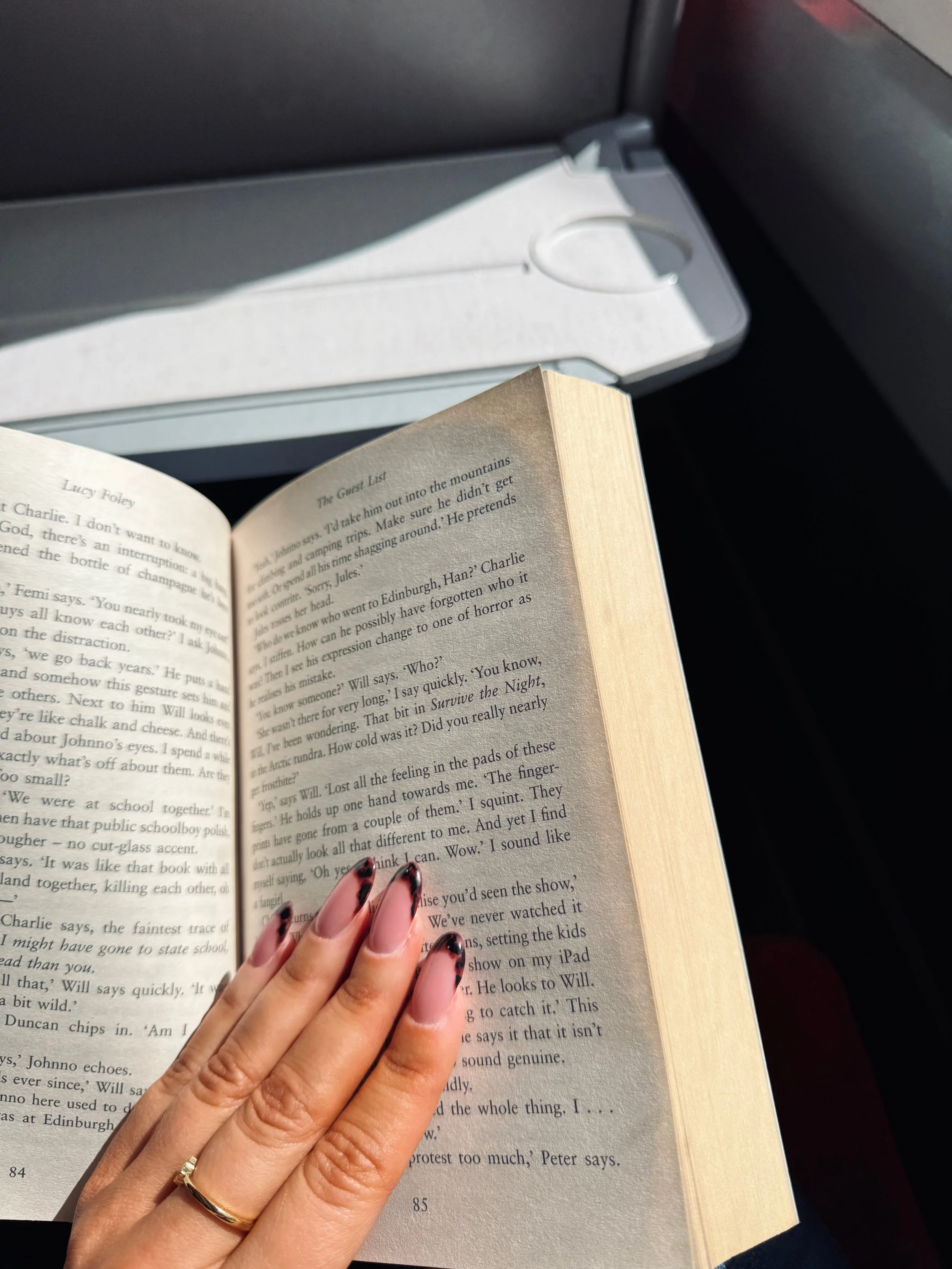A hand with manicured nails holds an open book, pages 84 and 85 visible, in sunlight. A tray table and seat on a train, appear in the background.