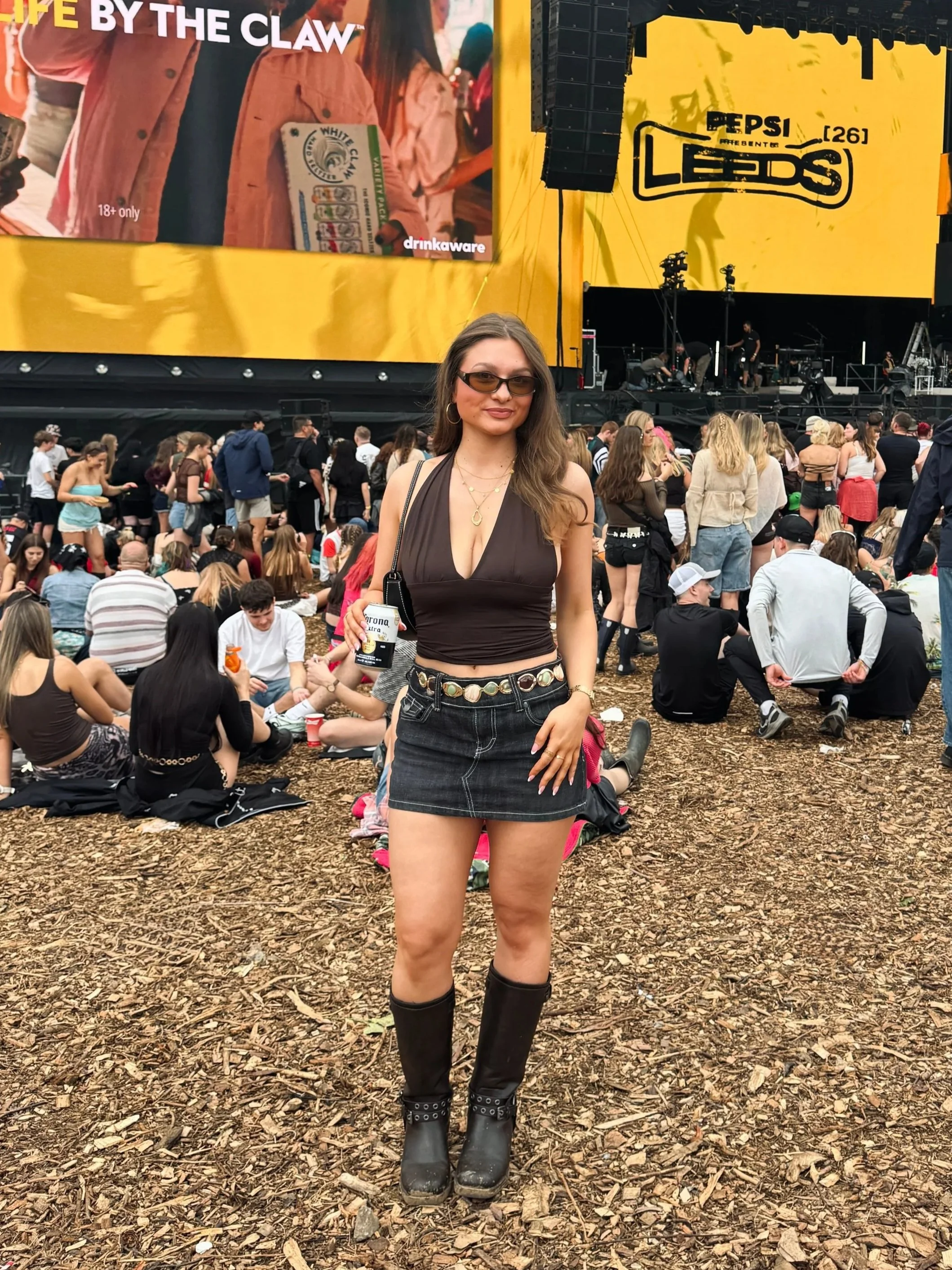 Woman in sunglasses, tank top, denim skirt, and boots poses at a lively concert, with a crowd, stage, and Pepsi screens behind her.