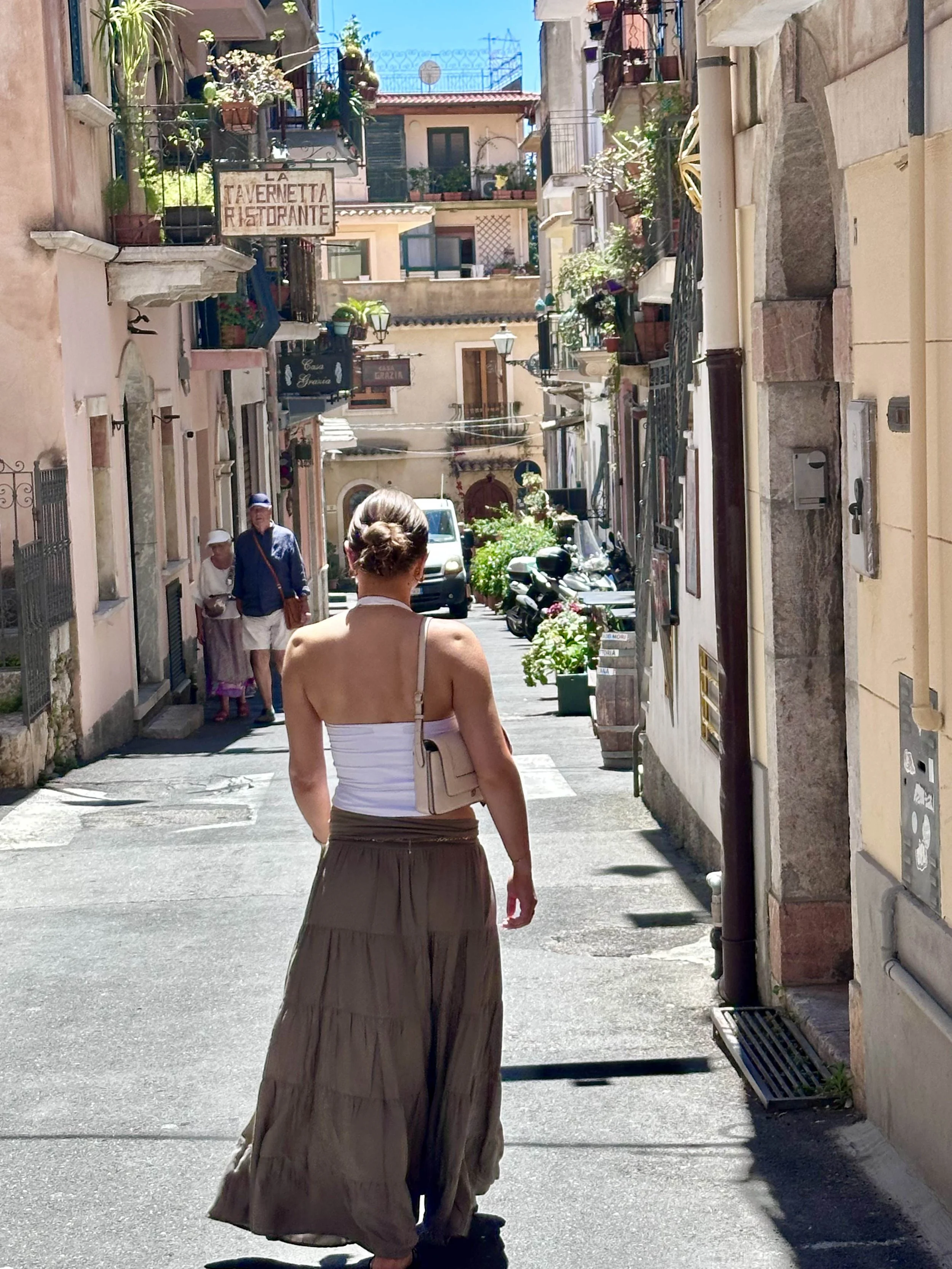 A woman walks down a narrow, sunlit street in a Mediterranean town, surrounded by charming buildings adorned with plants. A restaurant sign is visible in the background, adding to the picturesque setting