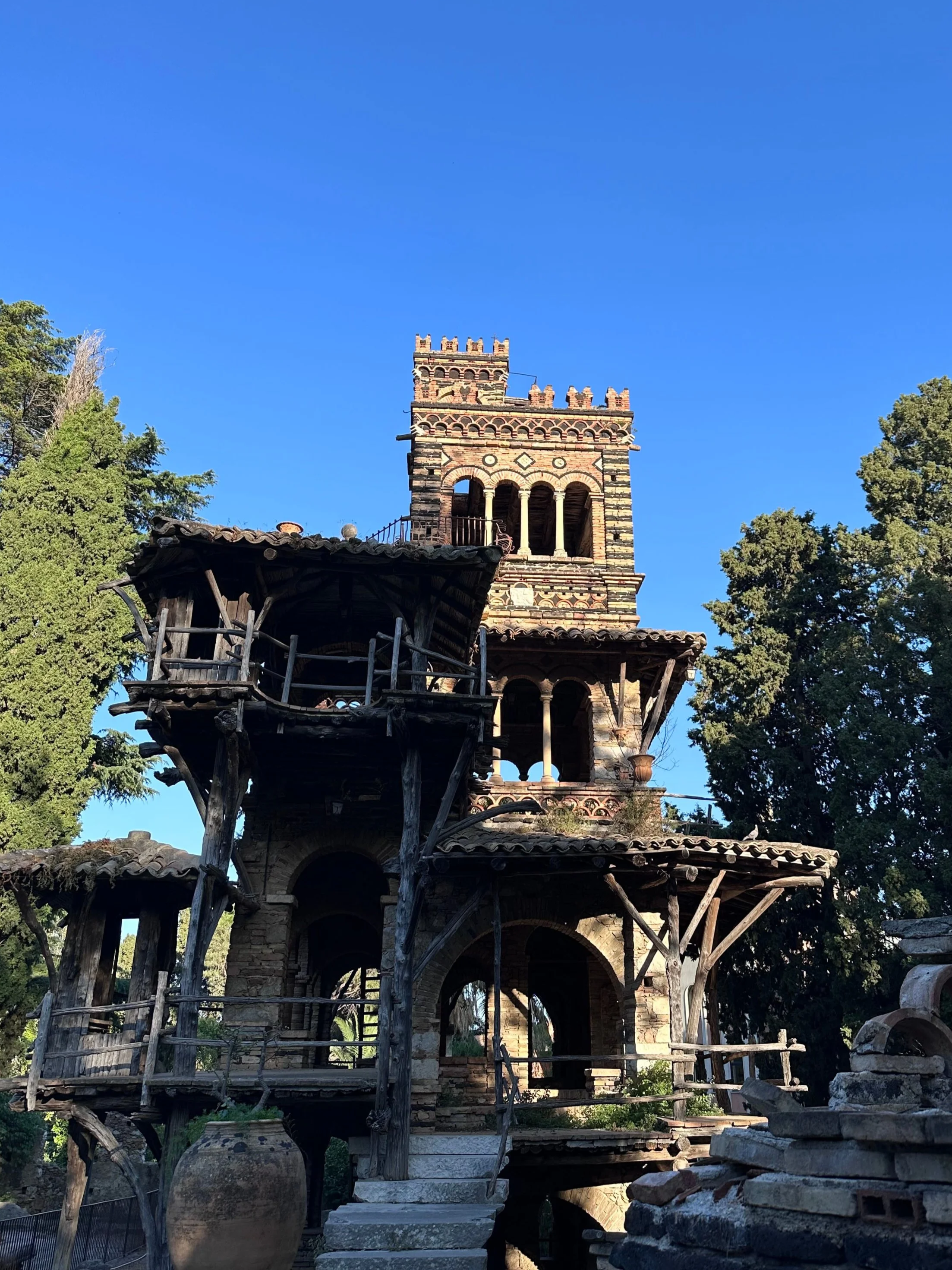 Rustic, multi-level stone tower with wooden balconies and arches, set against a clear blue sky and flanked by green trees, conveying a medieval ambiance.